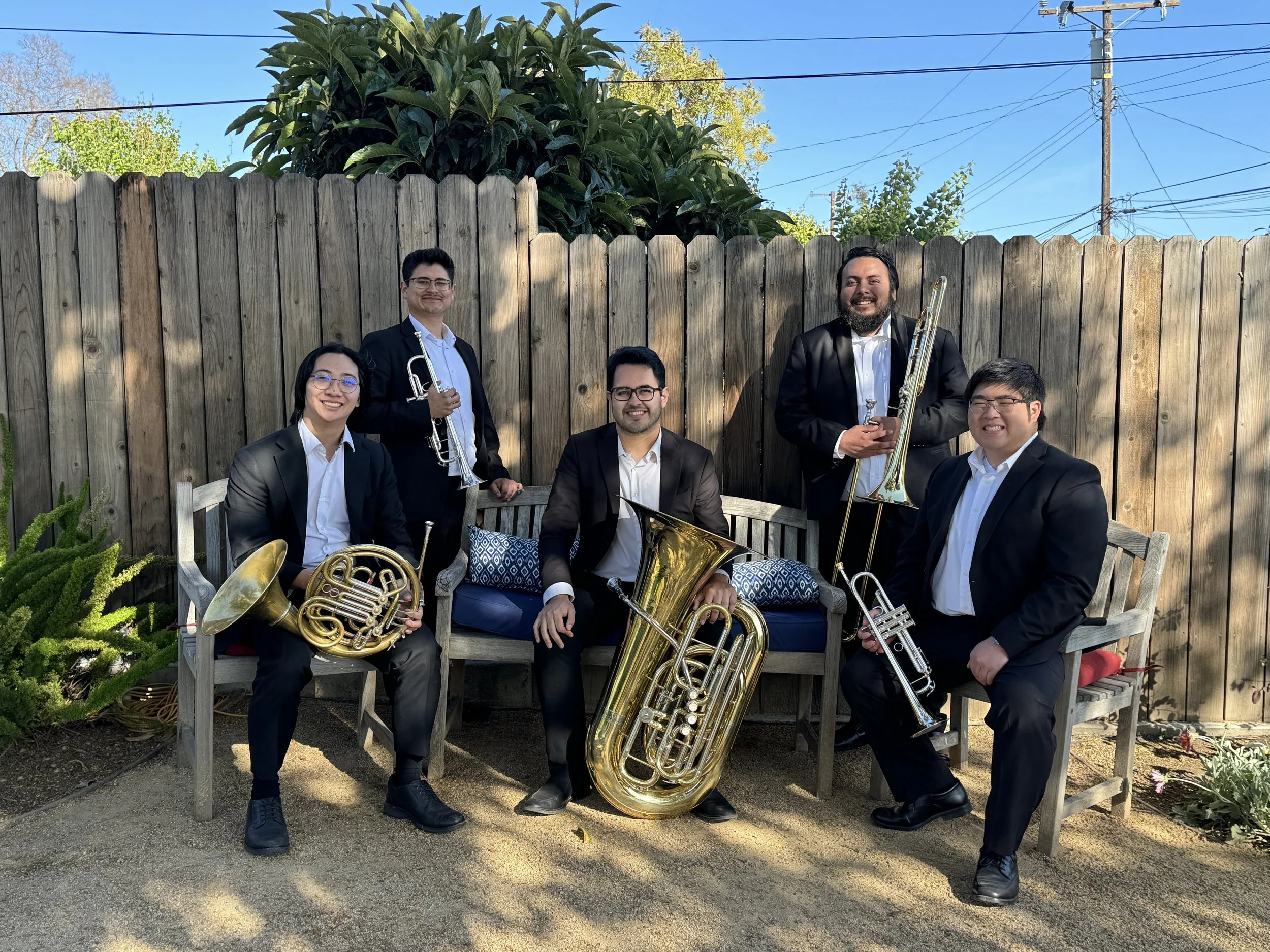 Five musicians dressed in formal black suits with white shirts, holding brass instruments, standing and sitting outdoors in front of a wooden fence with trees and power lines in the background.