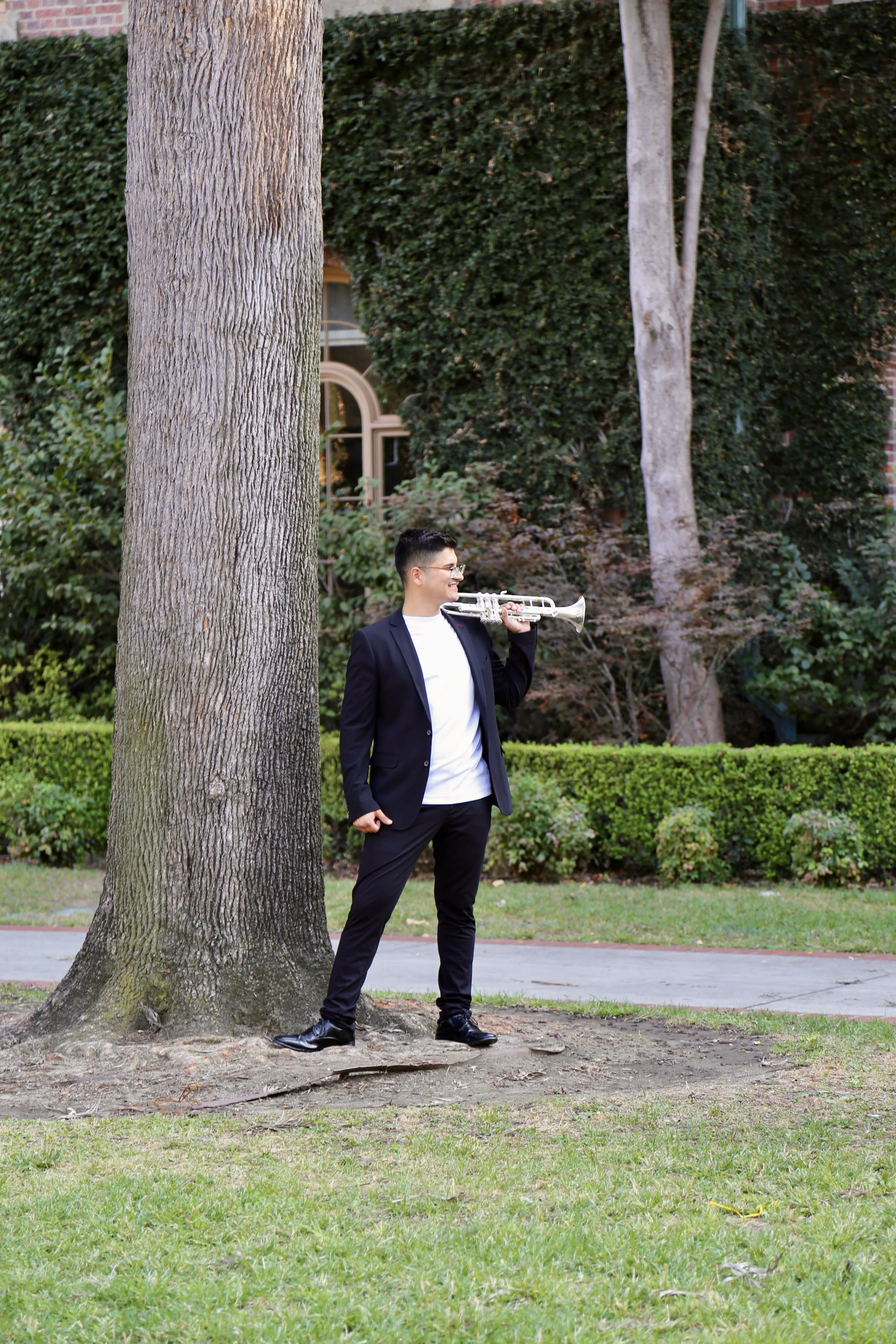 A man in a black suit and white shirt standing outdoors next to a large tree, smiling, holding a trumpet resting on his shoulder.