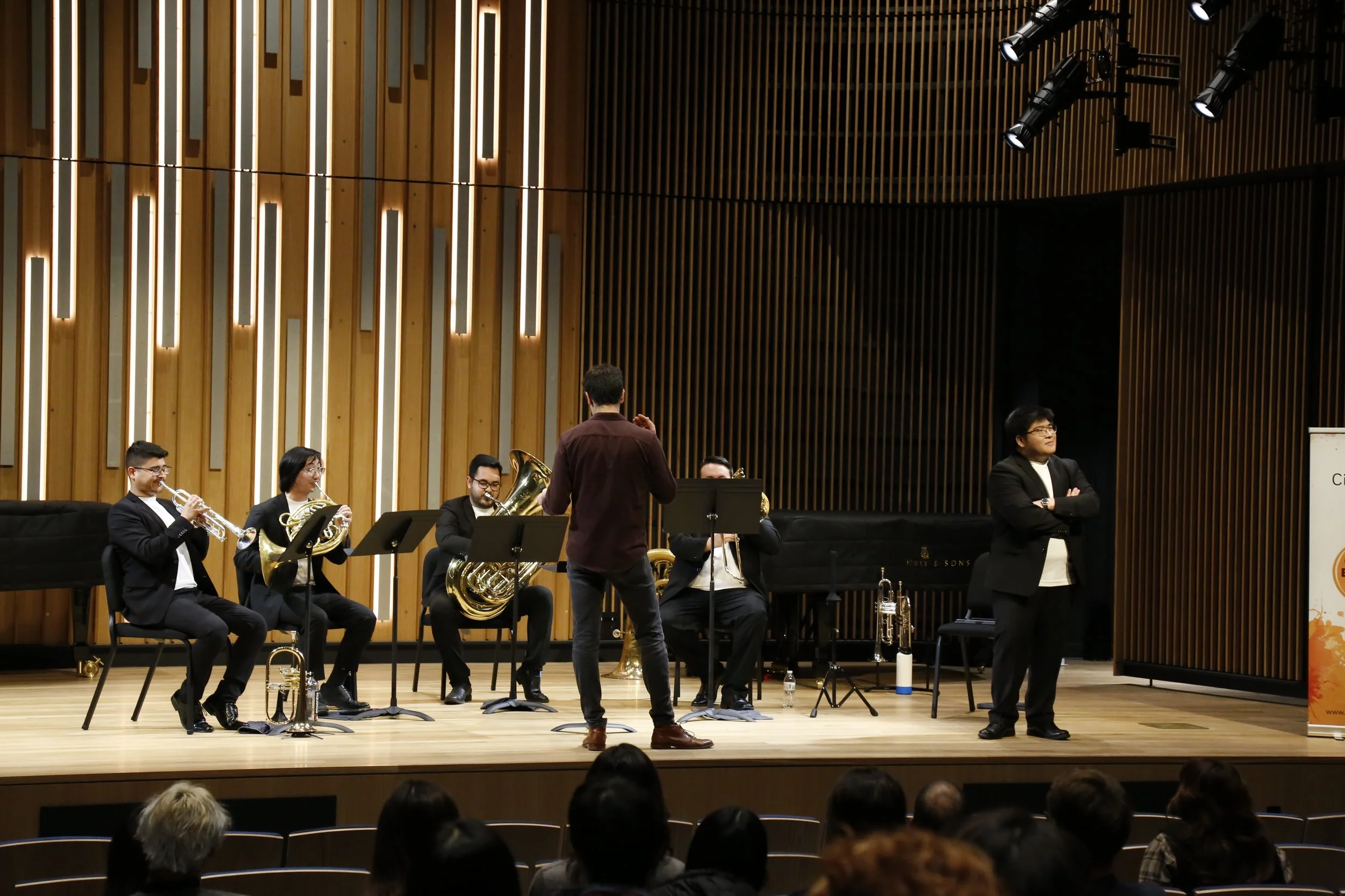 Musicians performing on stage during a concert, with a conductor facing them and an audience watching.
