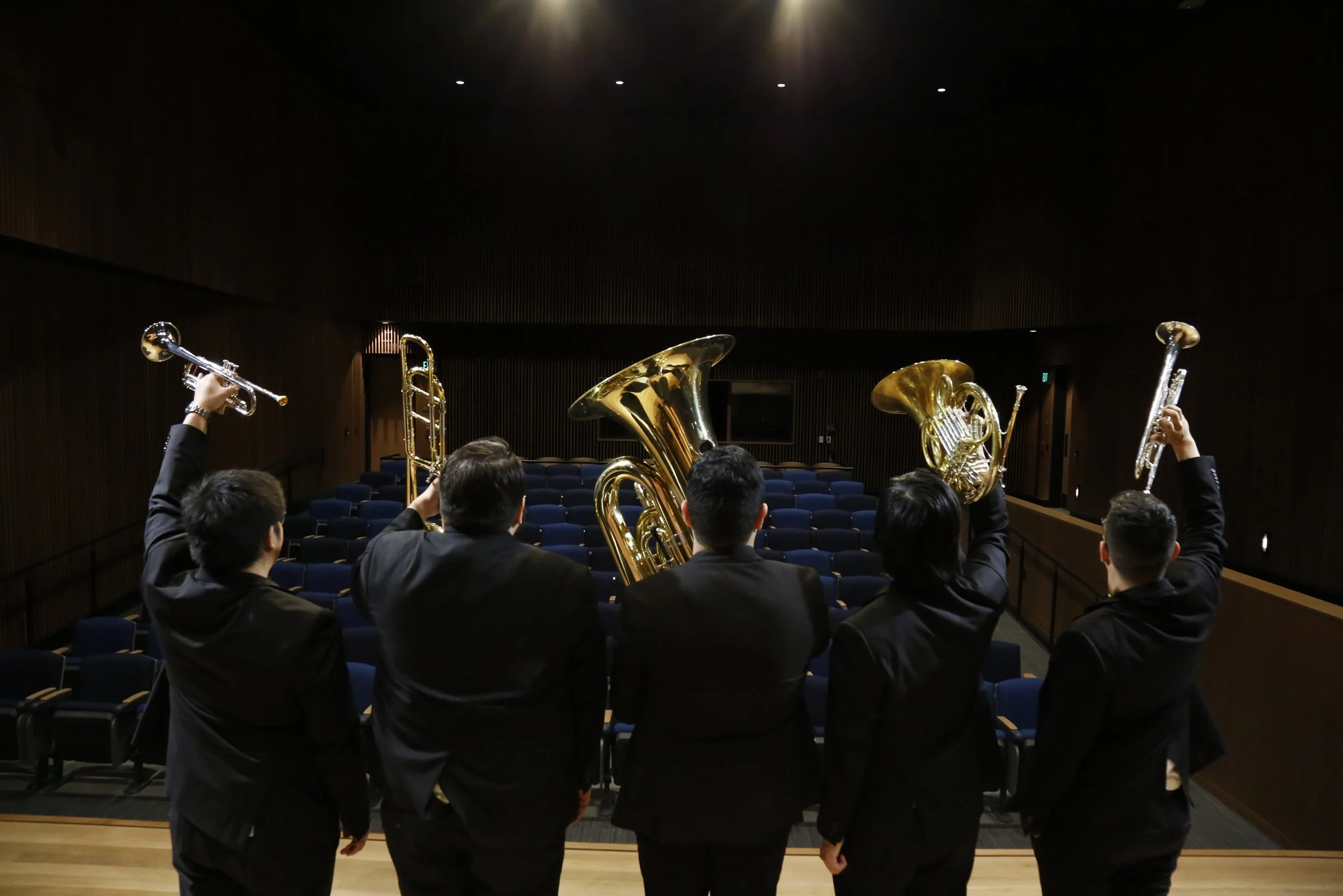 Group of five musicians in formal suits on stage holding brass and woodwind instruments in an empty auditorium.