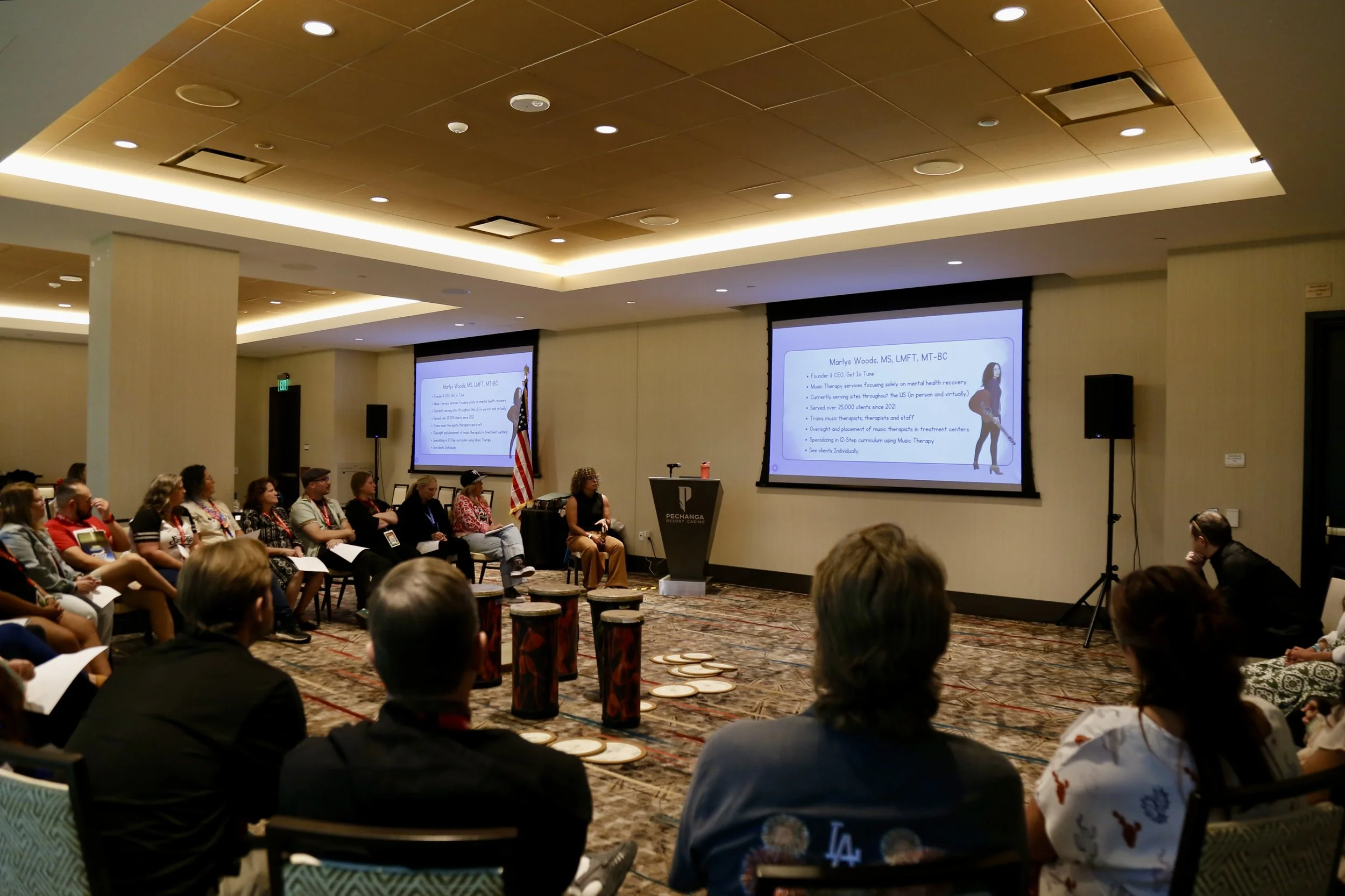 A professional conference room with attendees seated in a semi-circle, watching a presentation on two large screens. The presenter is a woman at a podium near an American flag. The room has a modern ceiling with recessed lighting and a patterned carp