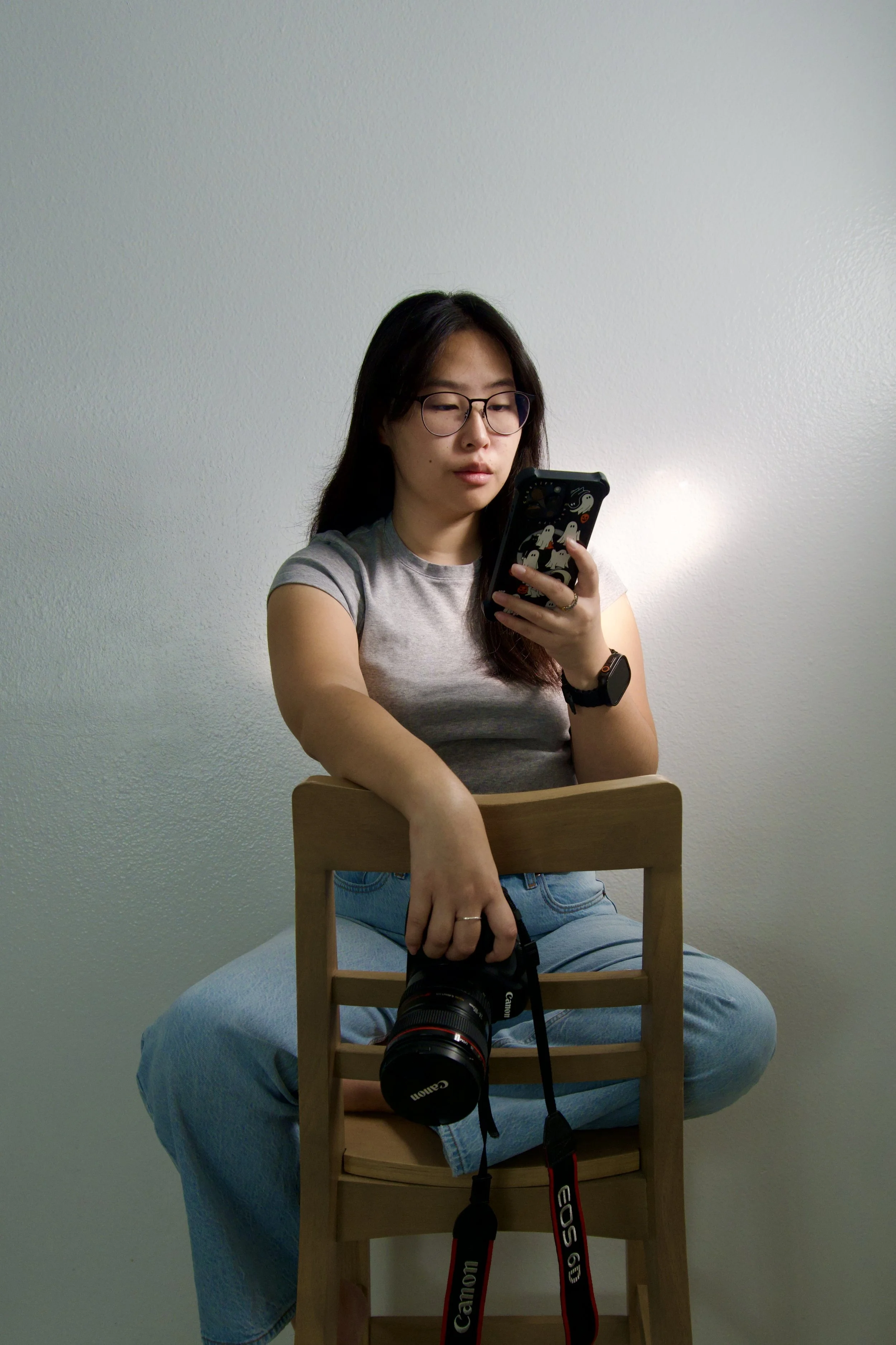 A woman sitting cross-legged on a chair, holding a camera in one hand and a smartphone in the other, with a plain wall in the background.