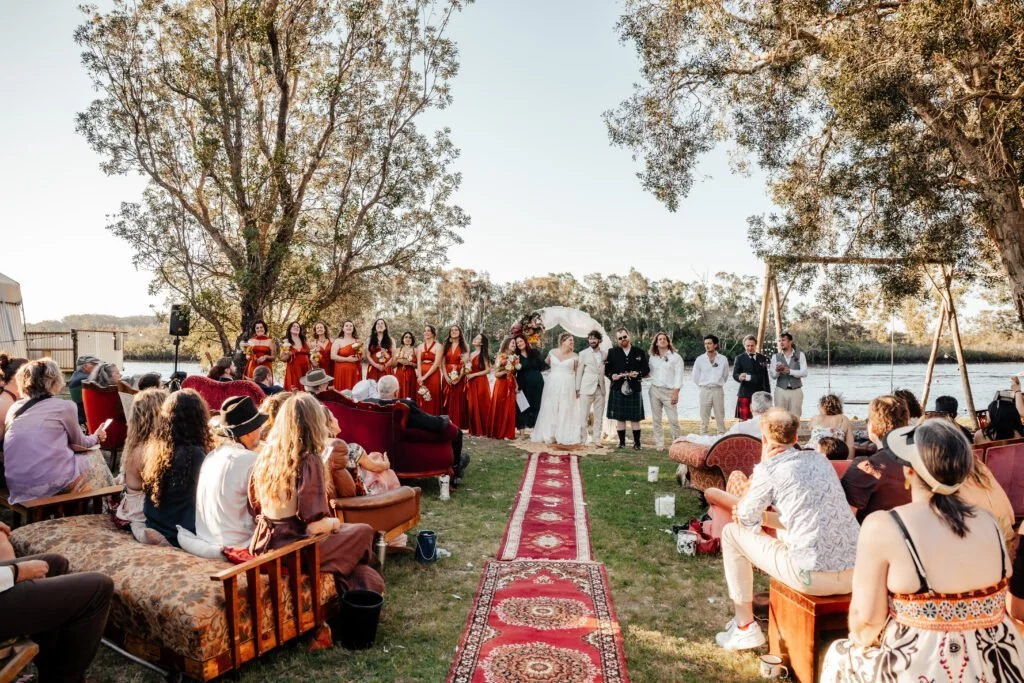 Outdoor wedding ceremony near a river at sunset, with the bride and groom kissing, surrounded by Leisure Disco Decor, bridesmaids in red dresses and groomsmen in white or gray, guests seated on sofas and chairs, trees and open sky in the background.