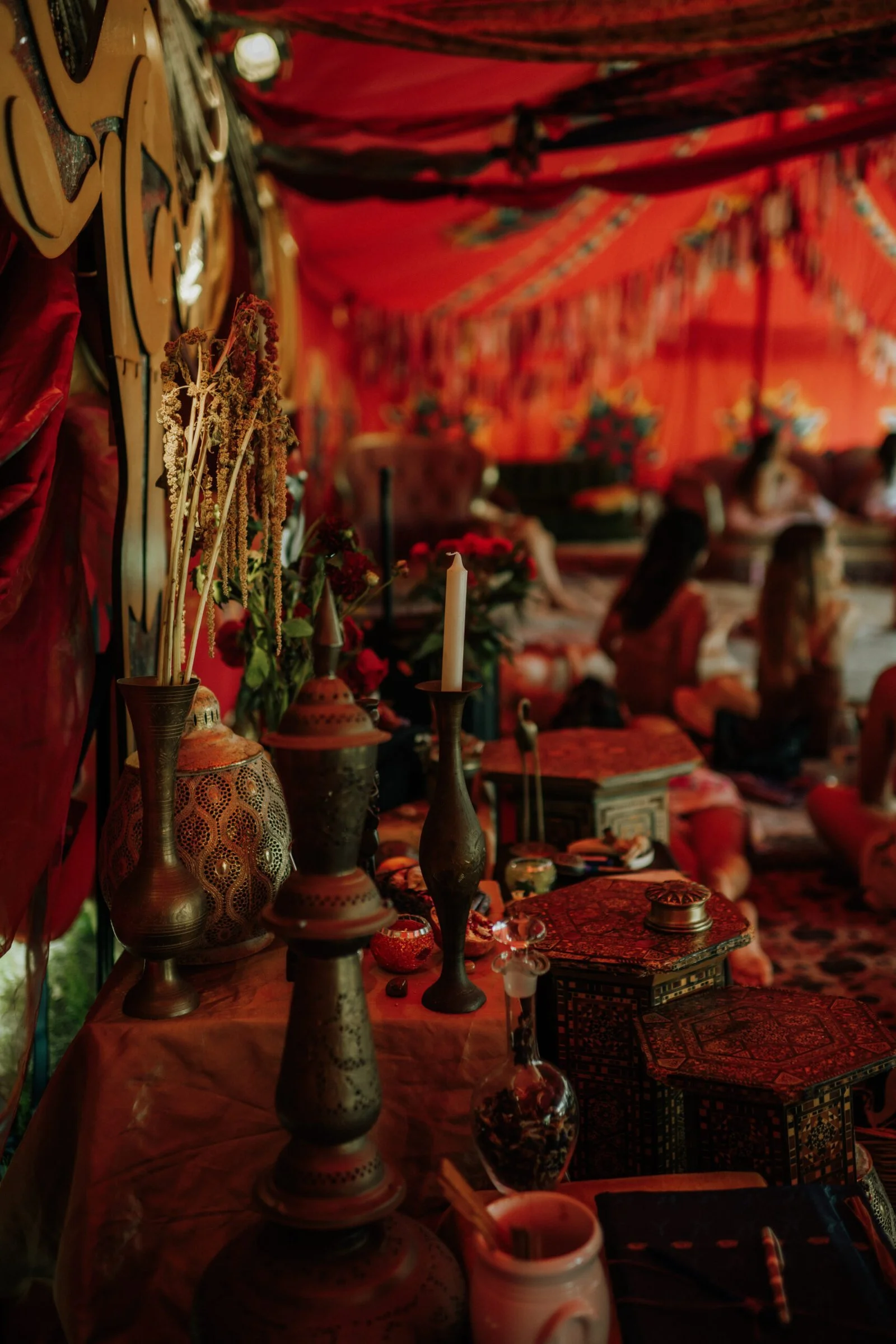 Decorative items such as candles, vases, and flowers on a table, with people sitting cross-legged on the floor in a decorated tent with vibrant red and orange fabric and patterns, suggesting a cultural or traditional setting.