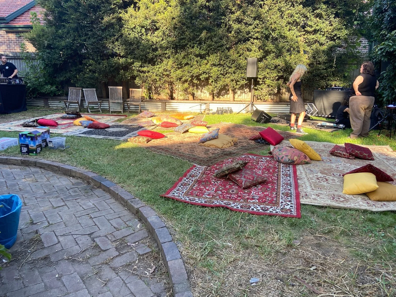 Outdoor backyard setup for a social event with patterned rugs and scattered cushions on grass, a DJ booth with a person behind it, and two women standing near music equipment, surrounded by trees and a fence.