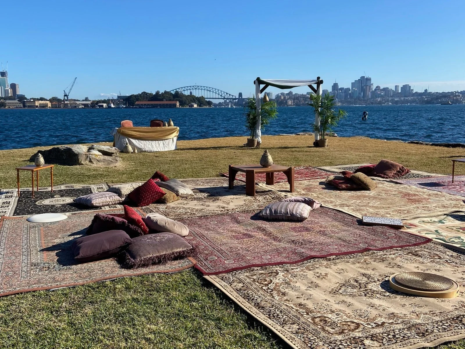 Leisure Disco Decor Outdoor picnic and wedding setup with Persian rugs and cushions on grass near water, with a trellis and potted plants, city skyline in the background.