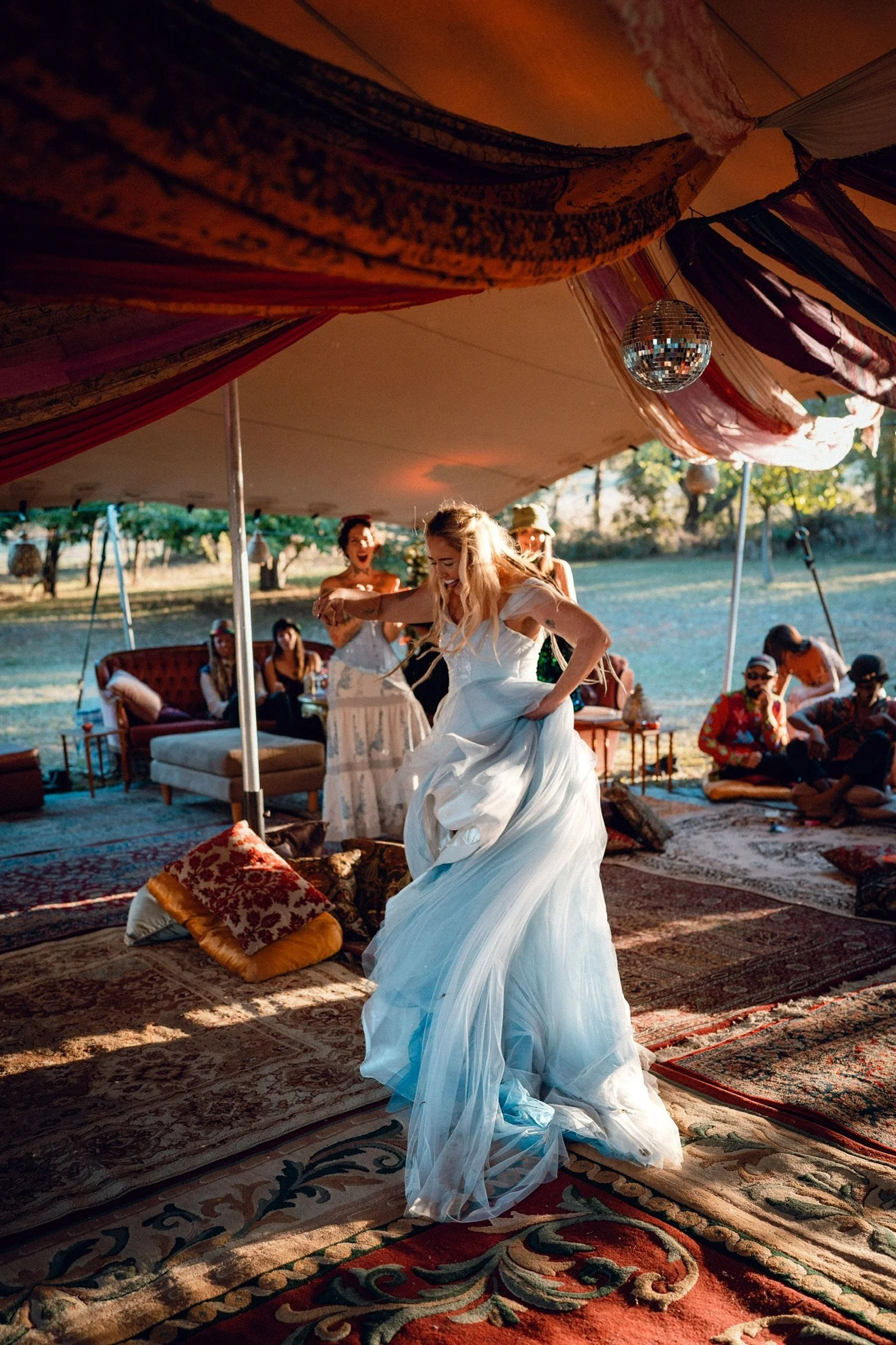 A bride in a white wedding dress dancing at an outdoor wedding celebration under a tent with friends sitting on cushions and rugs.