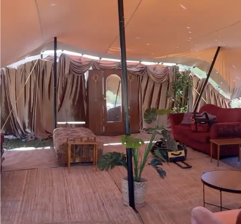 Interior of a tent with a red velvet sofa, potted plant, wooden tables, and draped curtains.