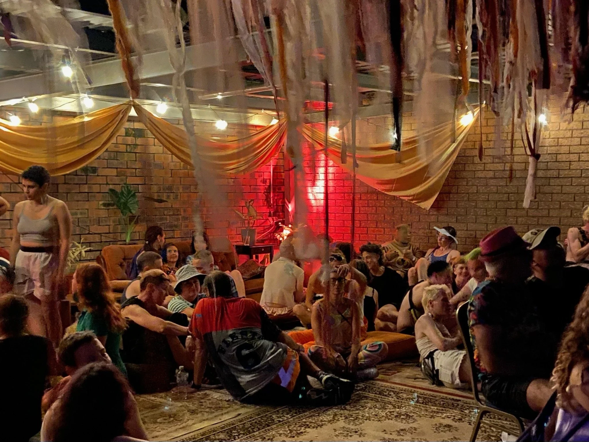 People gathered in a warmly lit indoor space with brick walls, draped fabric decorations, and ambient lighting, sitting on the floor and couches, engaging in conversations and socializing.