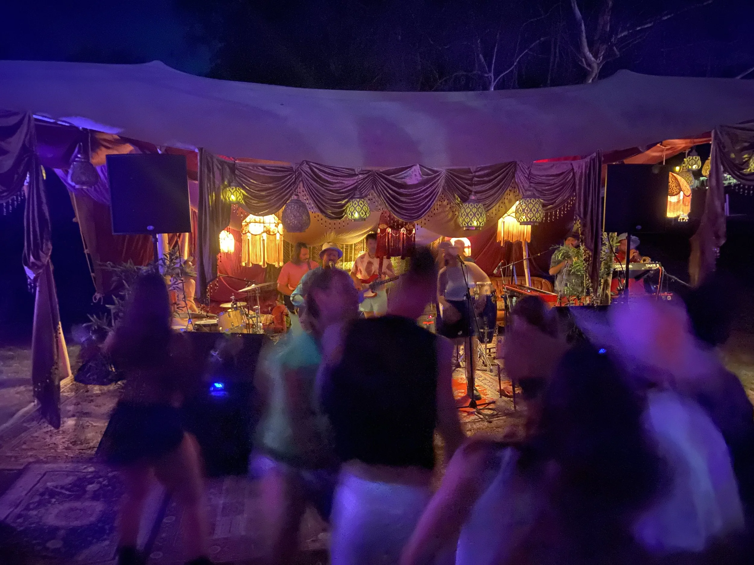 Musicians performing on an outdoor stage decorated with drapes and lanterns at night, with people dancing in front.