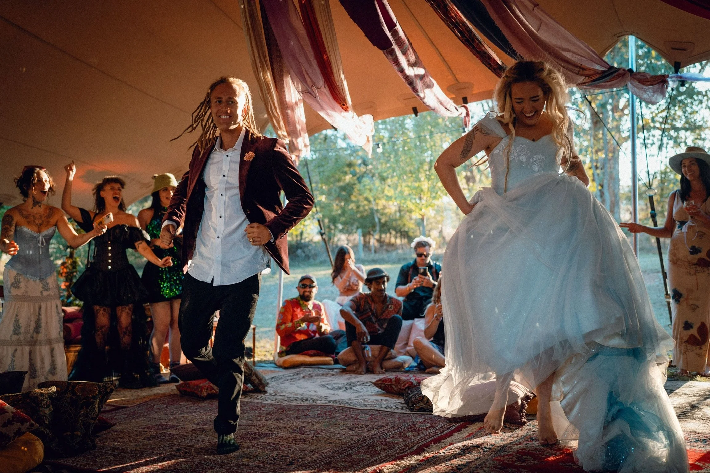 People dancing and celebrating at a wedding reception under a tent outdoors, with the bride in a white gown and the groom in a dark blazer, surrounded by guests. The scene is cheerful and lively with colorful decorations hanging overhead.