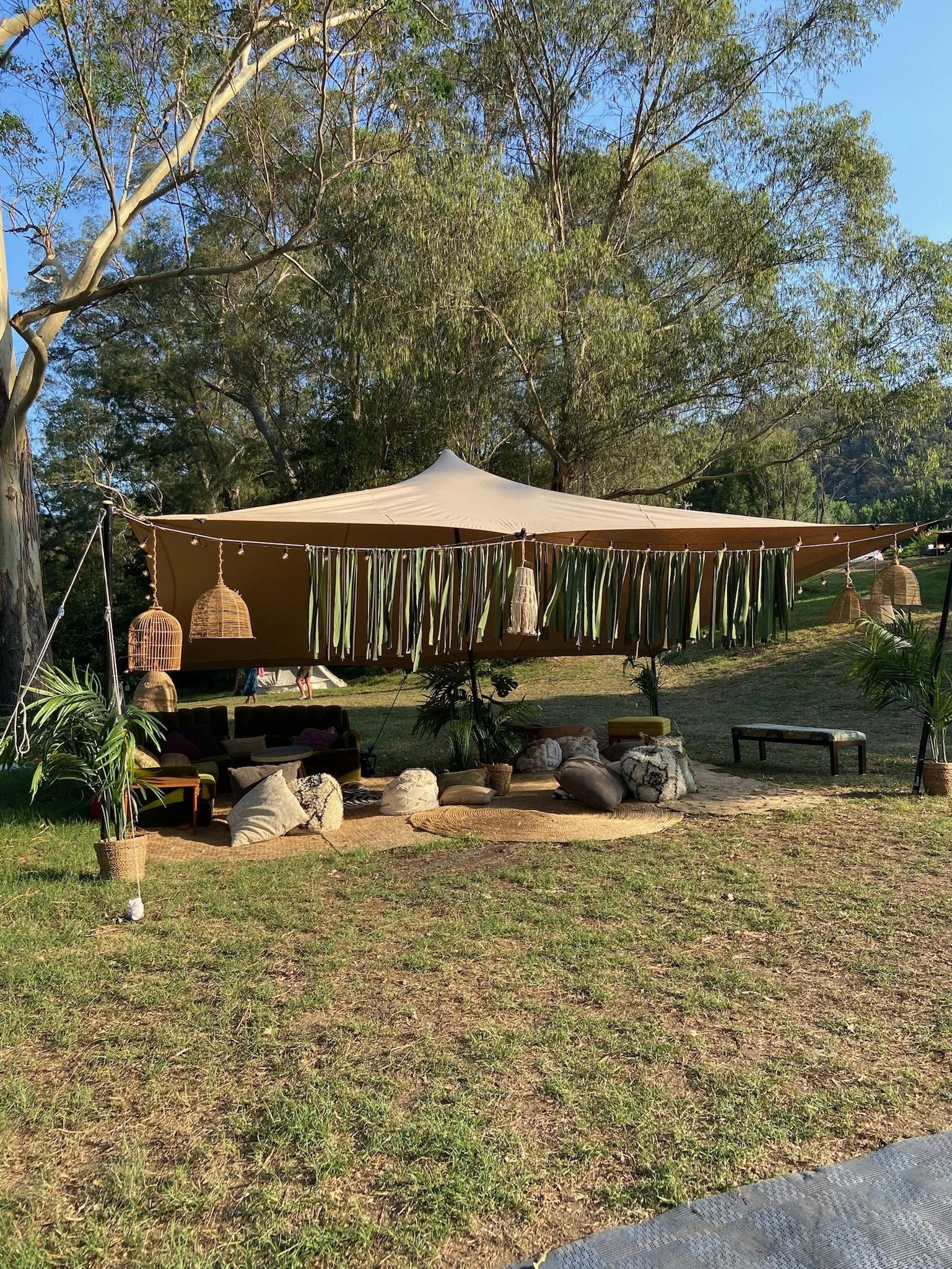 Outdoor gathering under a large tan canopy tent decorated with hanging lamps and fabric strips, with seating and cushions on the grass surrounded by trees and greenery.