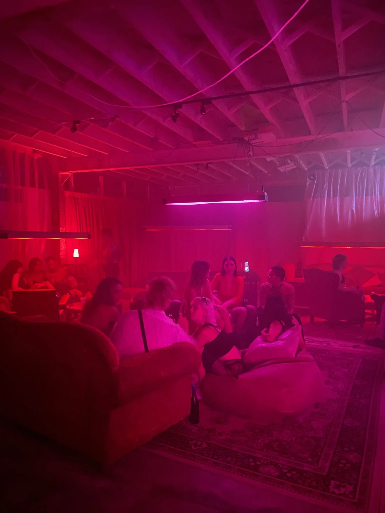 Group of people sitting on sofas and chairs in a dimly lit room with pink and purple neon lighting, engaging in conversation.
