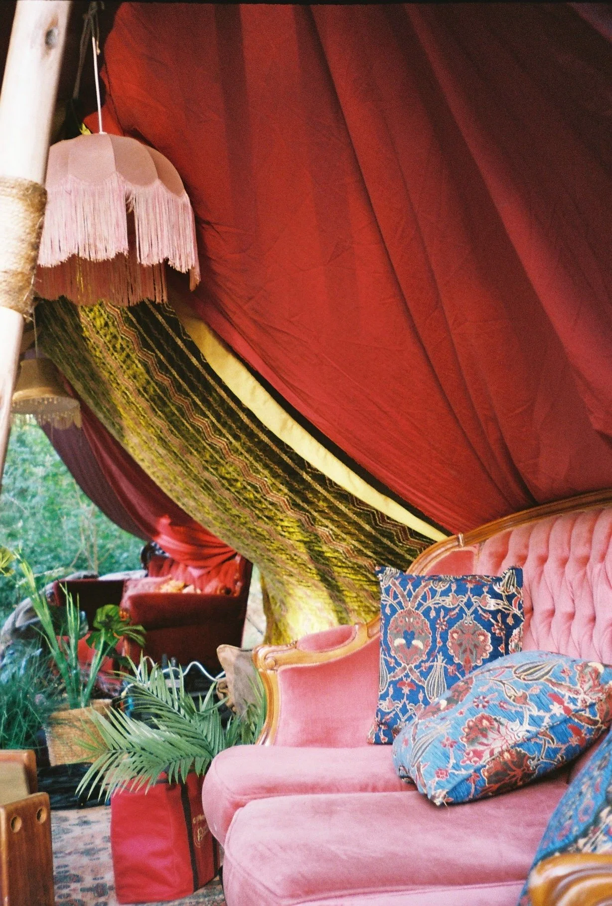 Interior of a room with vintage decor, including a pink upholstered sofa with decorative pillows, a side table with a fringed lampshade, lush green plants, a patterned curtain, and a booth with red upholstery in the background.