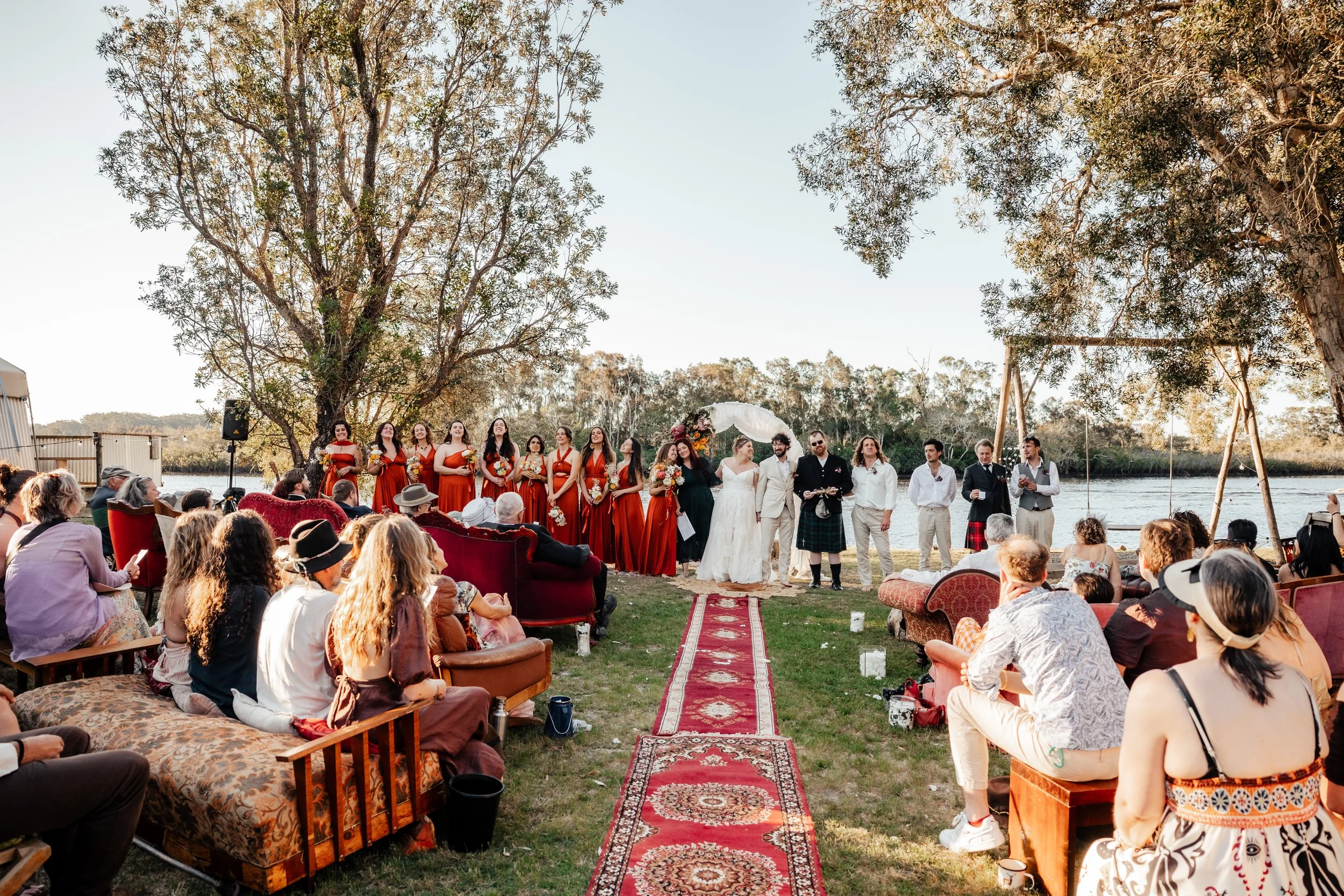 Outdoor wedding ceremony by a river with guests seated on sofas and benches, a bride and groom standing under a floral arch, and bridesmaids in red dresses, all surrounded by trees and an open sky.