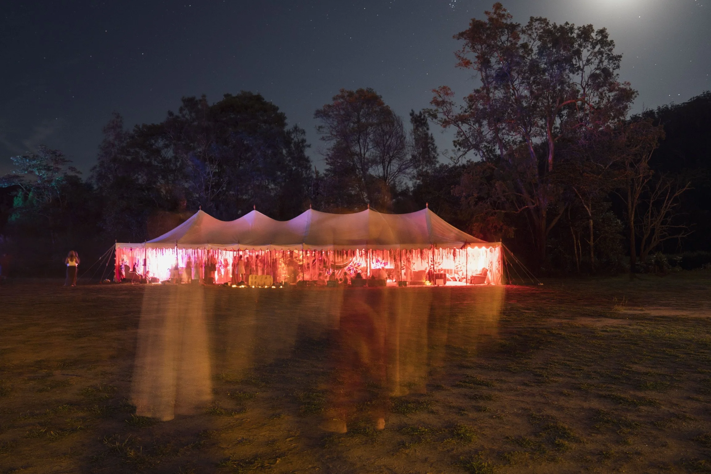 A large, illuminated tent at night with colorful lights, positioned on a grassy field with trees in the background and a starry sky overhead.