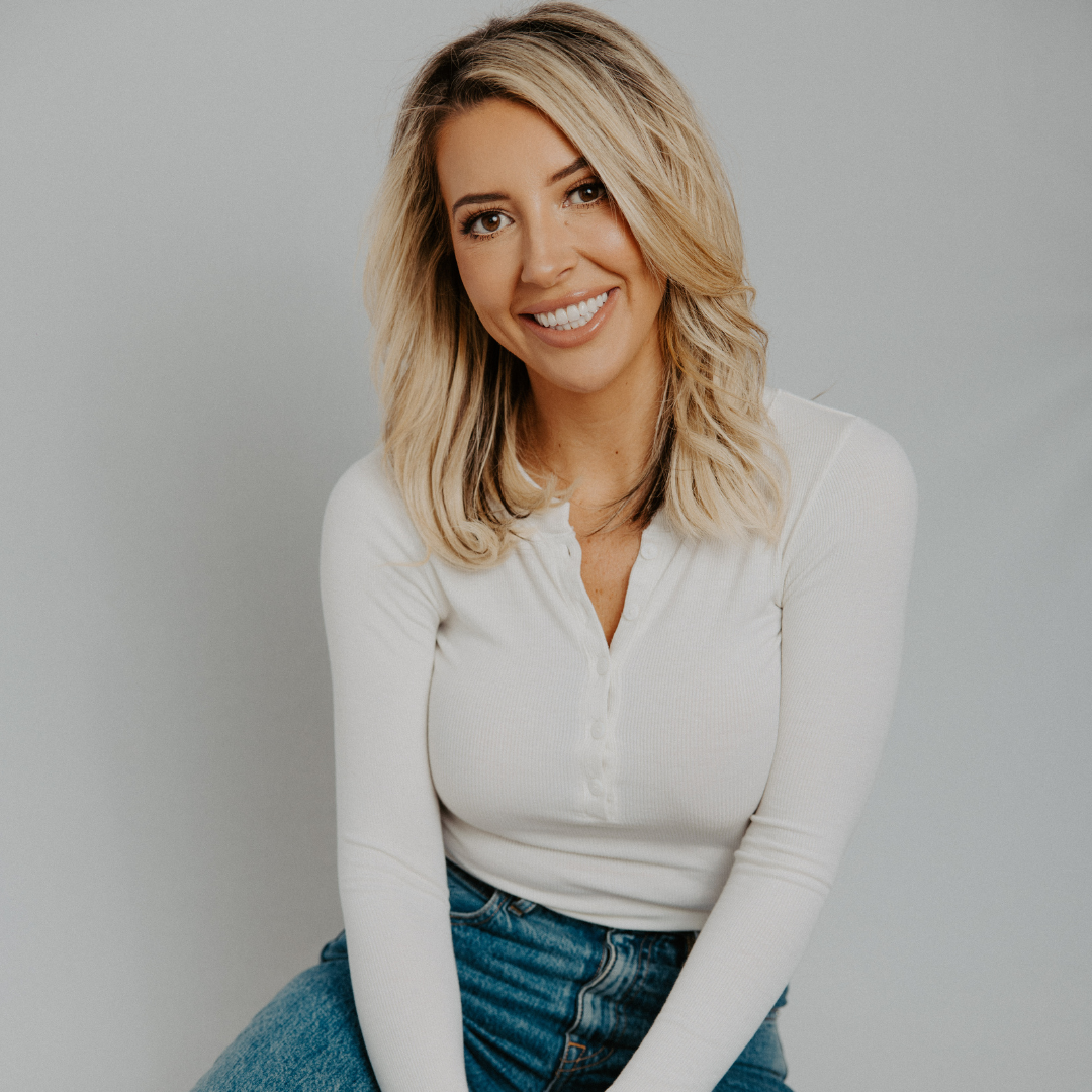 A woman with blonde hair smiling and winking, wearing a white long-sleeve shirt and blue jeans against a plain light gray background.