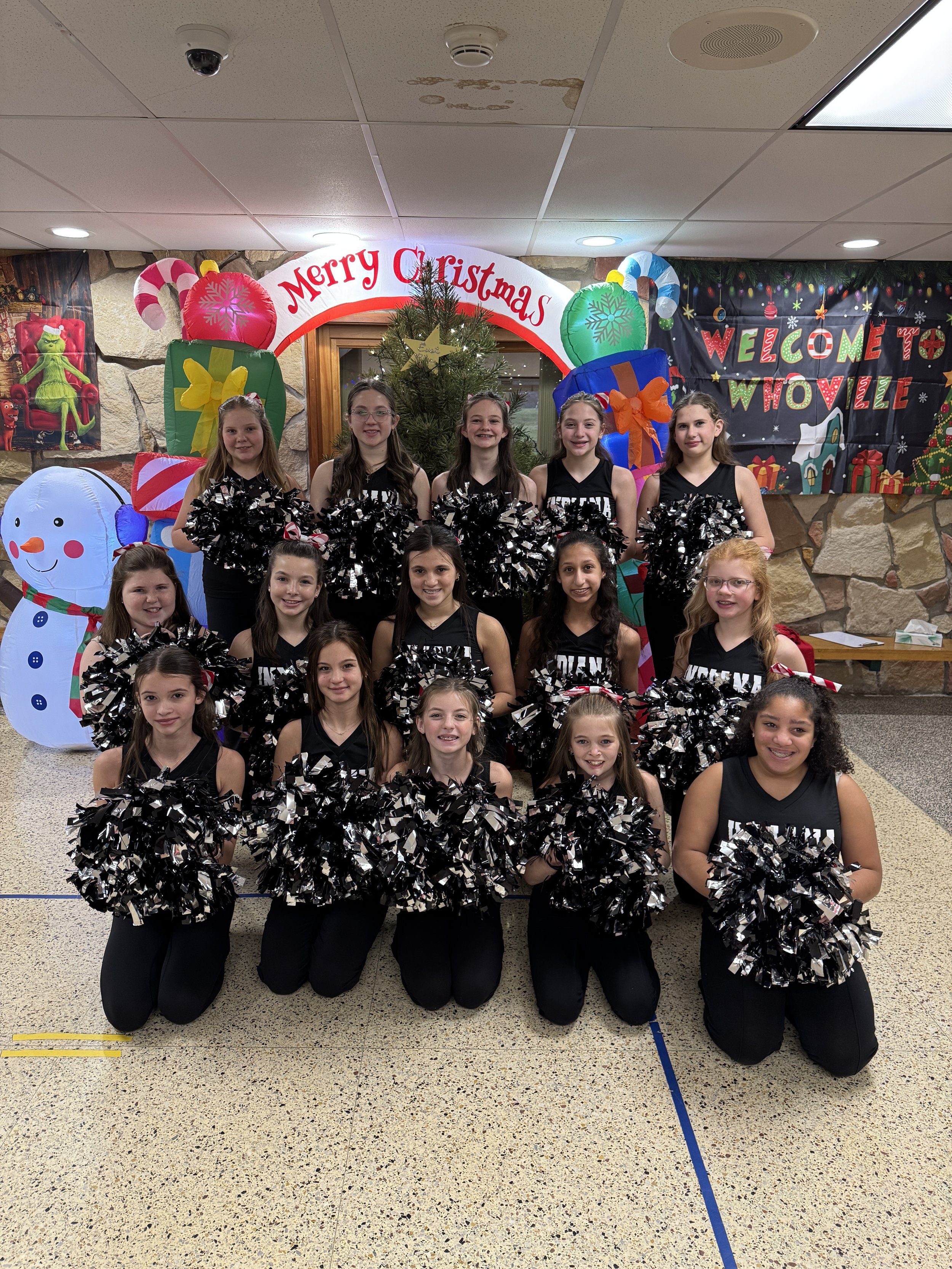 Group of young dancers wearing red and black uniforms with silver and black and red pom-poms, kneeling and standing in front of a Christmas decorated background with a Christmas tree, snowman, and banners that read "Merry Christmas".