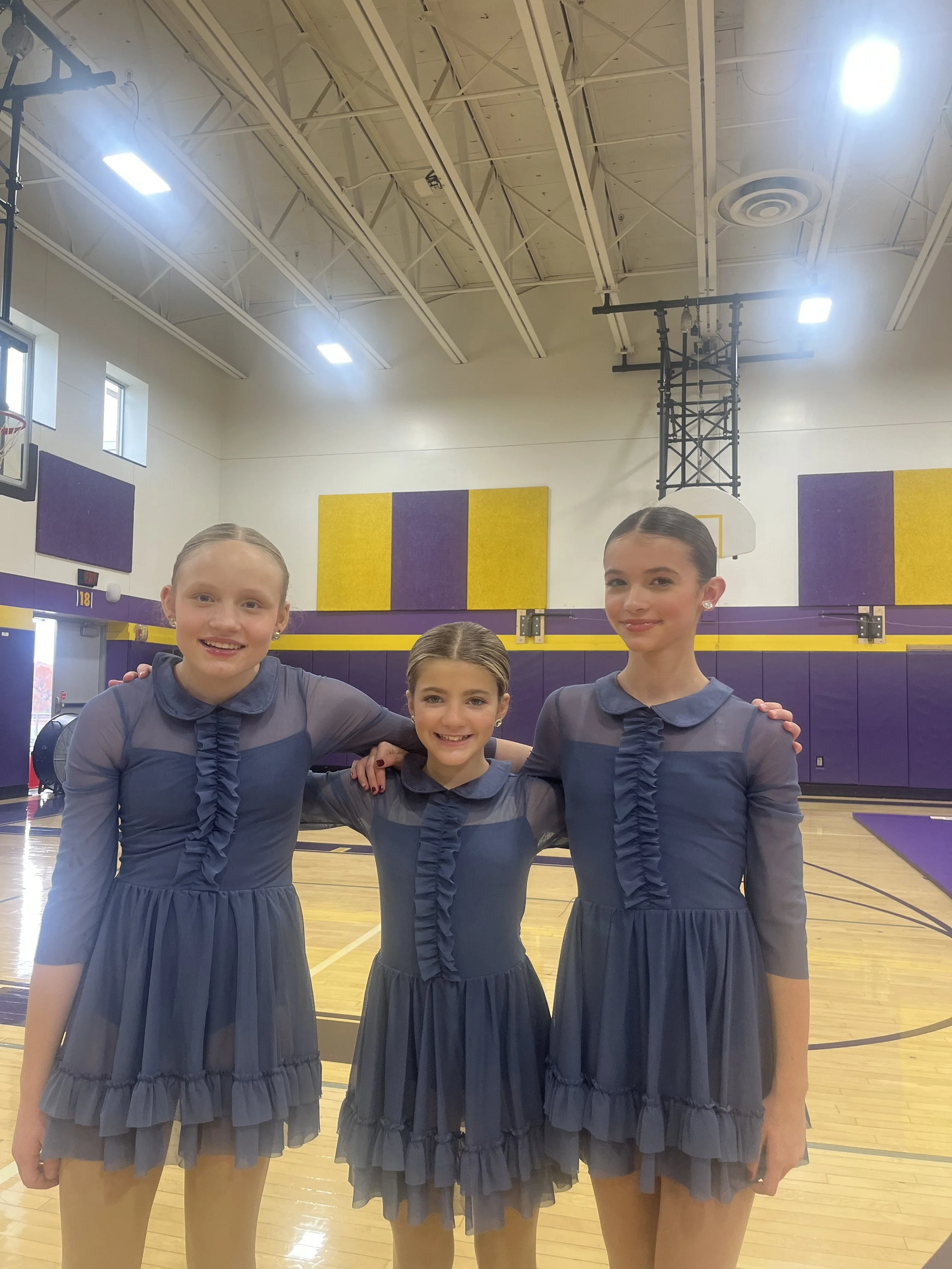 Three young girls dressed in matching blue costumes standing arm-in-arm in a gymnasium with purple and yellow walls, hardwood floor, and basketball hoop in the background for a dance competition.