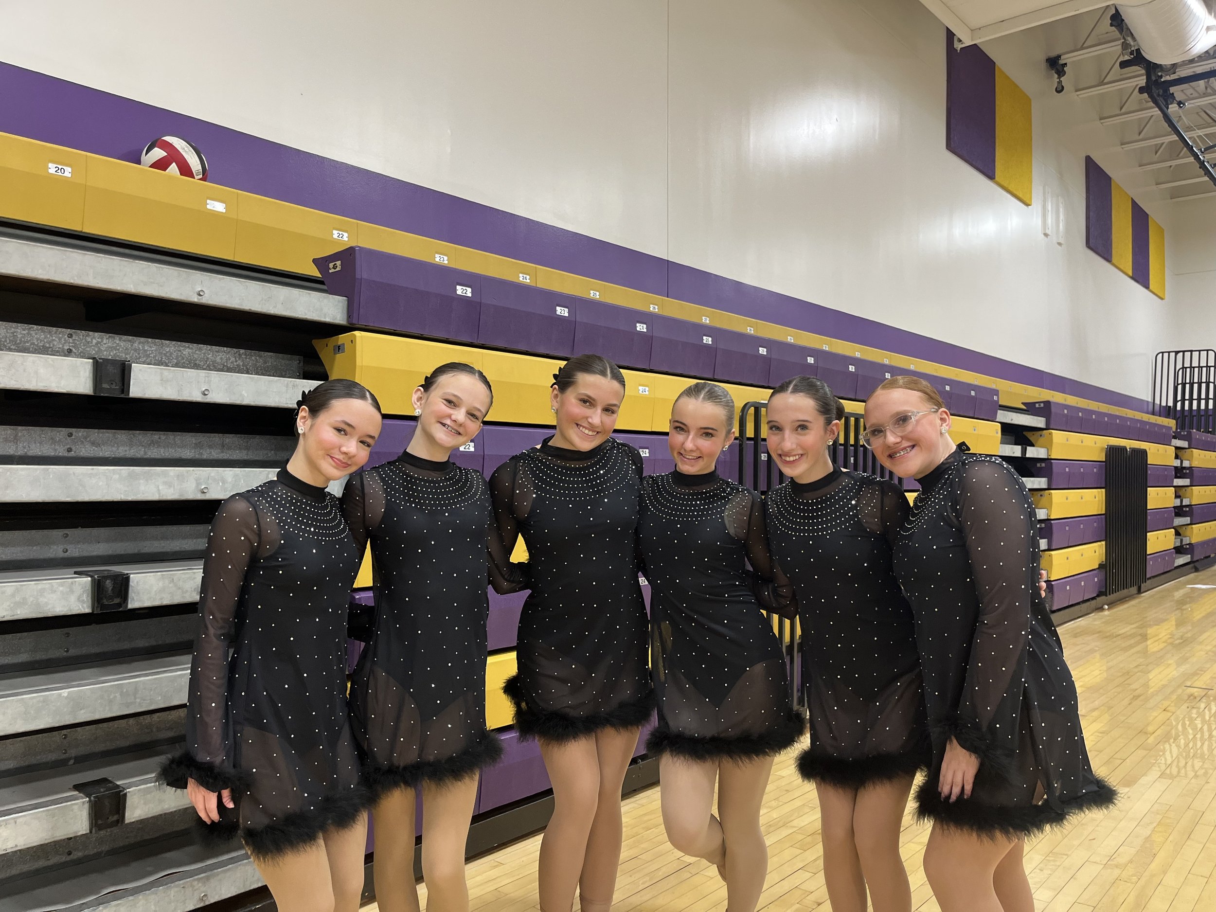Six young women in matching black costumes with rhinestones and feather trim, standing inside a gymnasium with purple and yellow bleachers for a dance competition.
