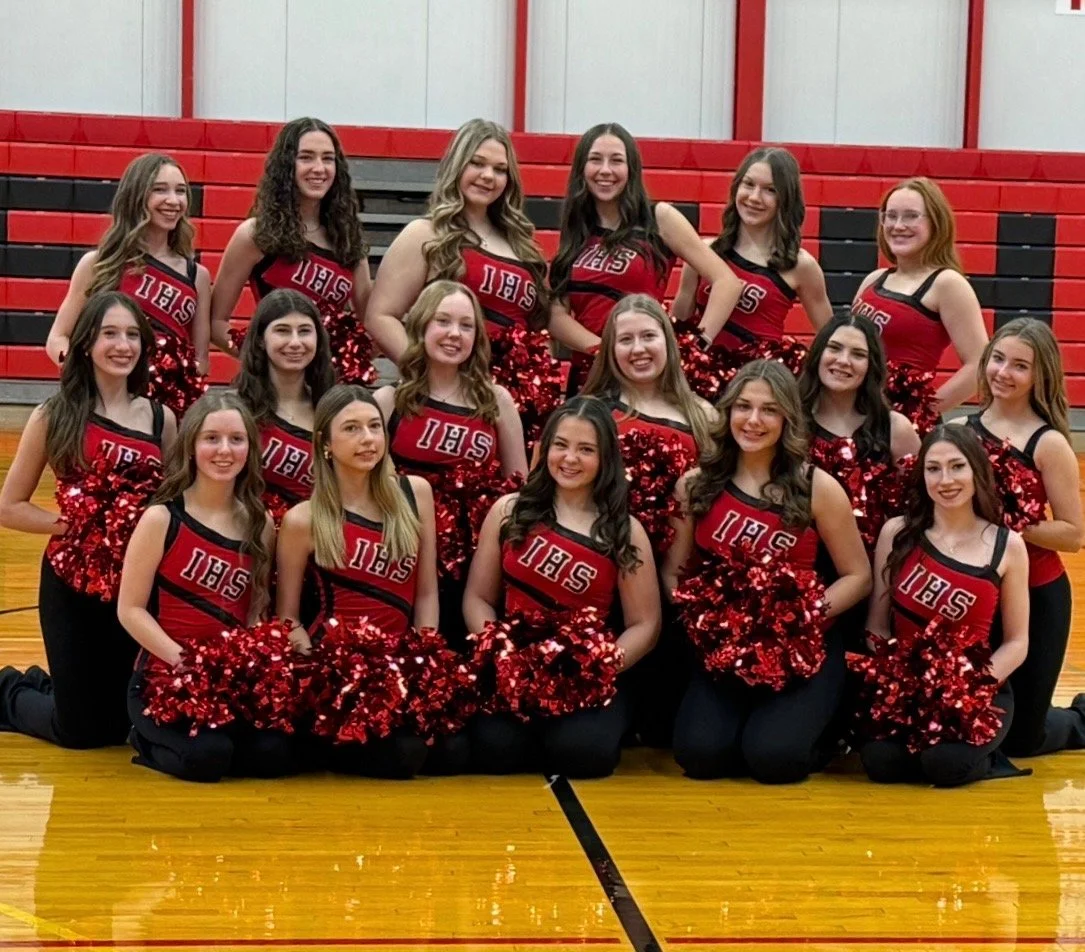 A group of dancers in red and black uniforms with 'IHS' on the front, holding red pom-poms, kneeling and standing on a basketball court in a gymnasium with red bleachers behind them.