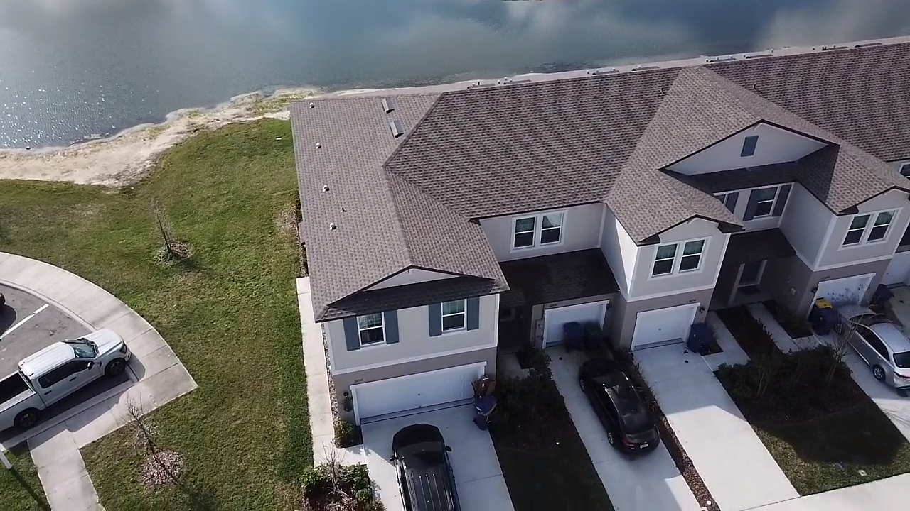 Aerial view of a row of townhouses with attached garages, parked cars, green lawns, and a body of water in the background.