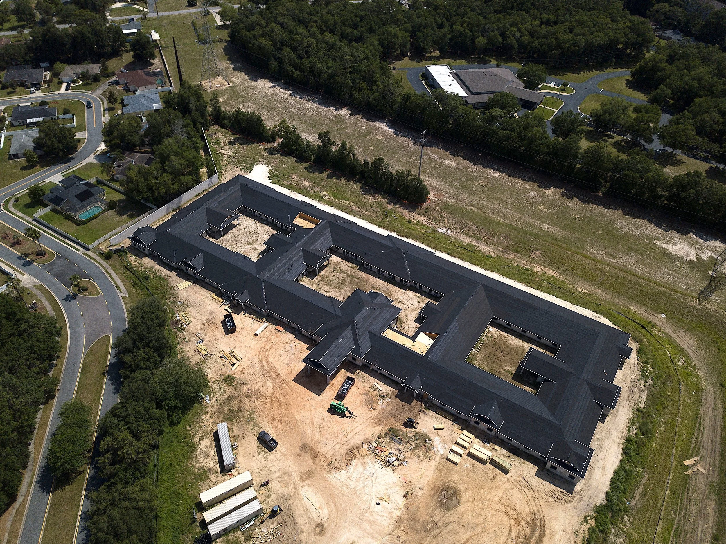 Aerial view of a large construction site while a new roof is being installed, surrounded by dirt and construction equipment, with residential homes, trees, and roads nearby.