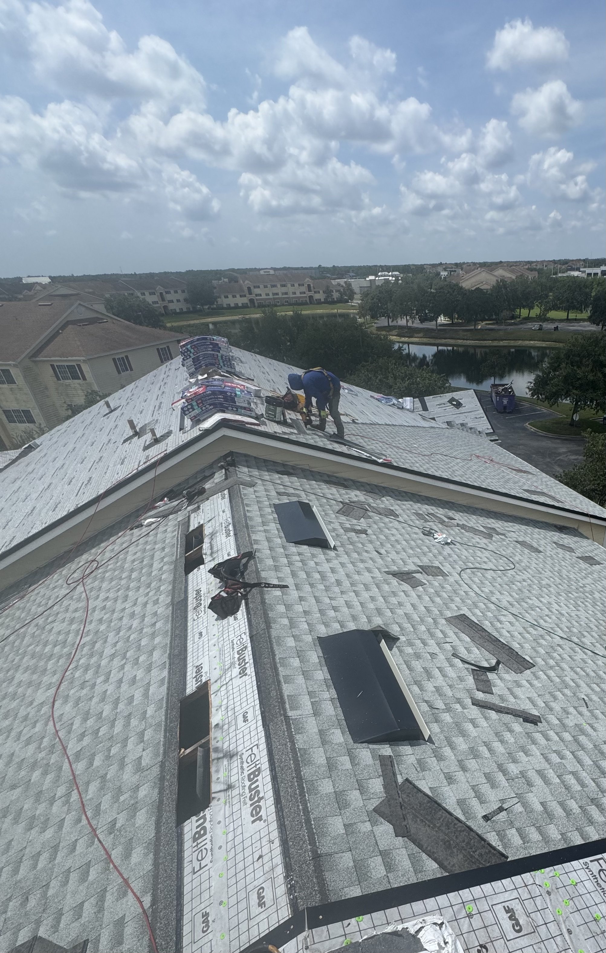 Two construction workers installing roofing on a house, with roofing materials and tools scattered on the roof, overlooking a residential neighborhood, some trees, a pond, and a cloudy sky.