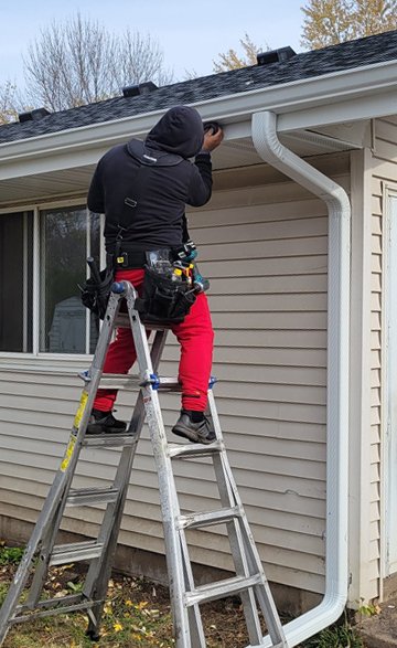 Worker on a ladder repairing or inspecting a house's rain gutter in an outdoor setting.