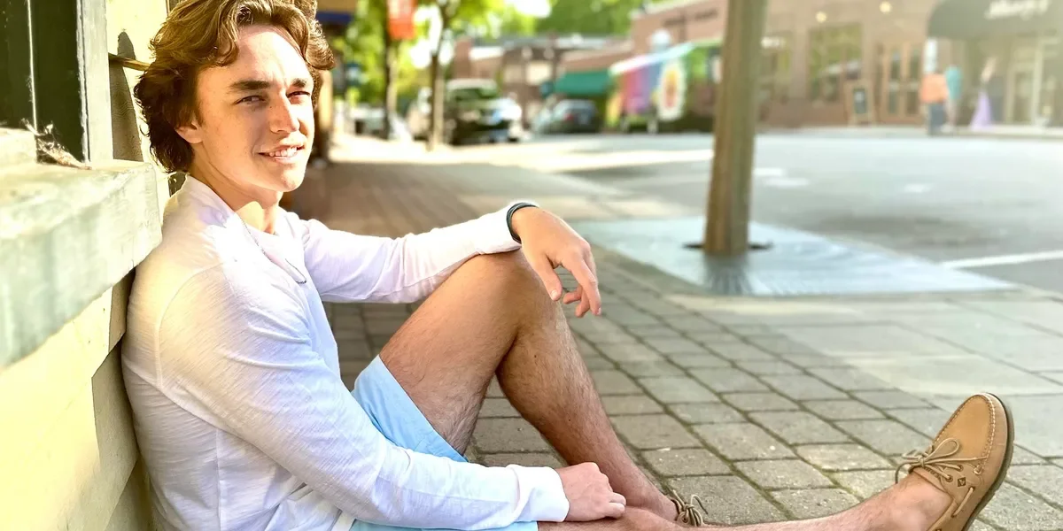 A young man with wavy brown hair sitting on a sidewalk, leaning against a yellow wall, wearing a white long-sleeve shirt, blue shorts, and tan boat shoes, smiling at the camera.