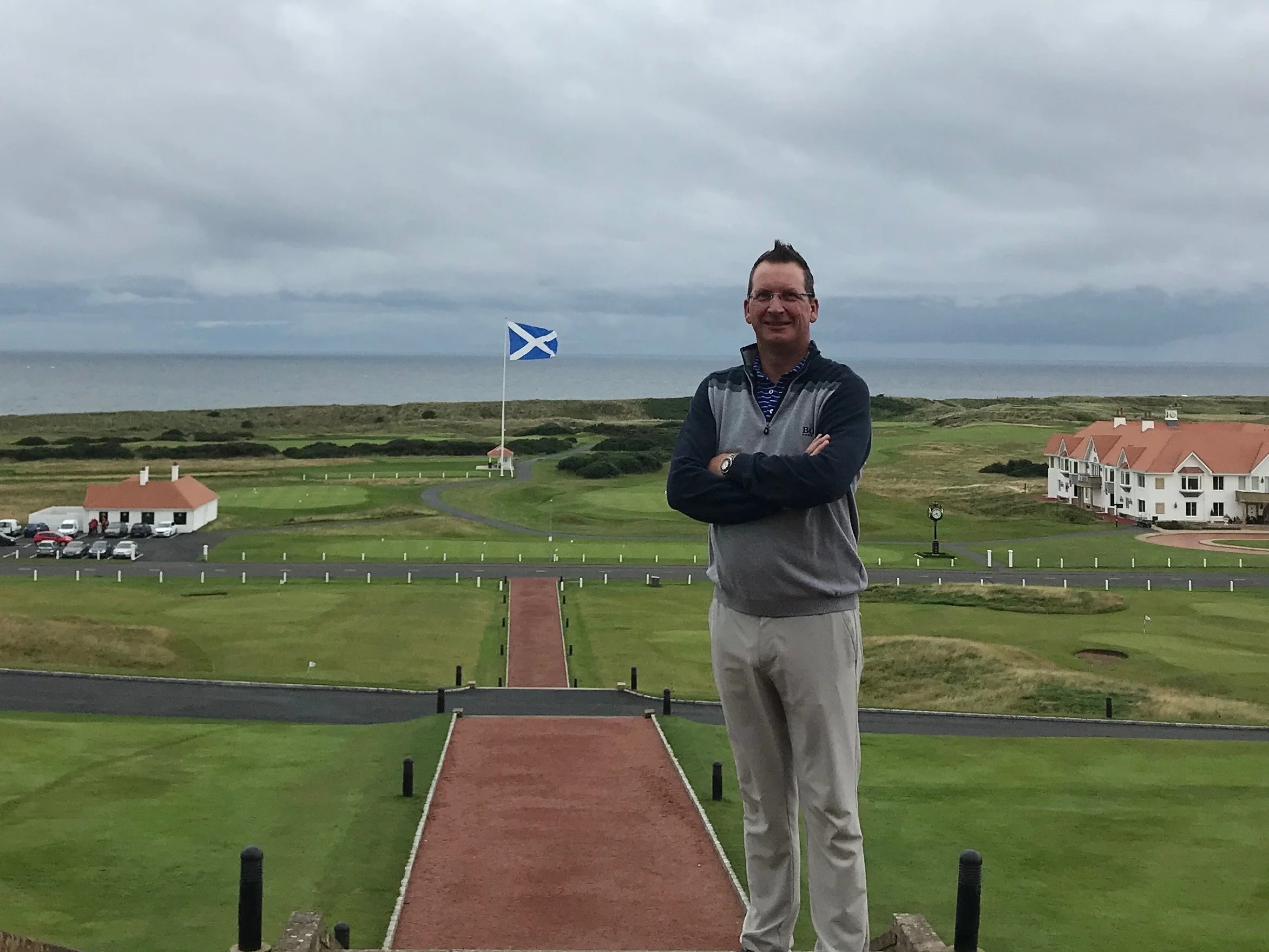 A man standing on a golf course with cross arms, smiling, near the coastline, with a Scottish flag in the background, under cloudy skies.