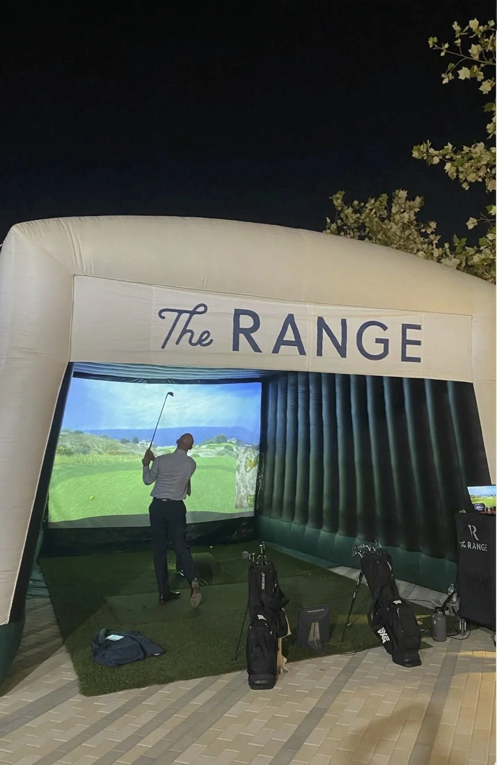 An indoor golf simulator setup labeled 'The Range' with a person swinging a golf club, surrounded by golf bags and equipment, inside a tent-like structure.