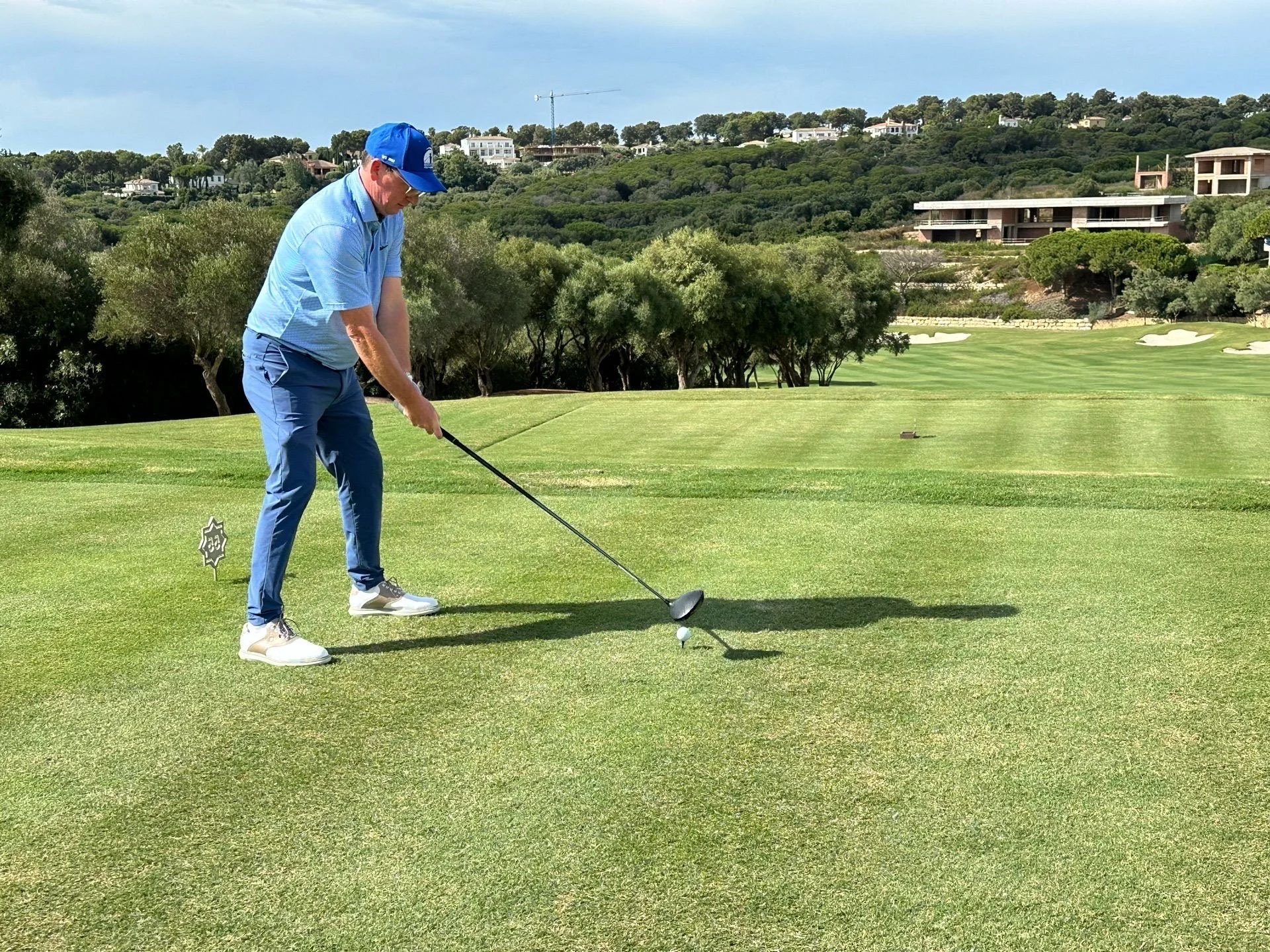 A man wearing a blue shirt, blue pants, and a blue cap is preparing to hit a golf ball on a golf course with a driver club.