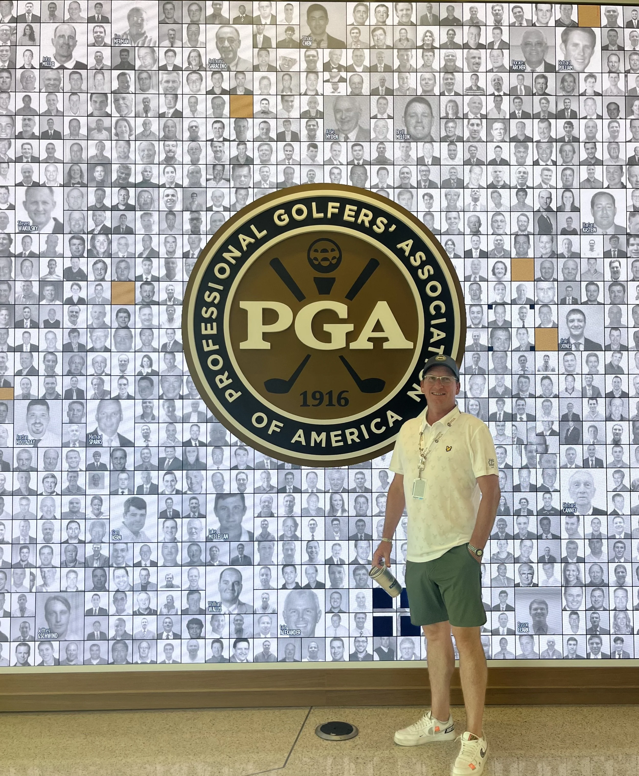 A smiling man wearing a baseball cap, glasses, a white polo shirt, green shorts, and Nike sneakers standing in front of a large wall with a collage of black and white photos of many people. The wall features a large gold and black PGA logo for the Professional Golfers' Association of America, established in 1916.