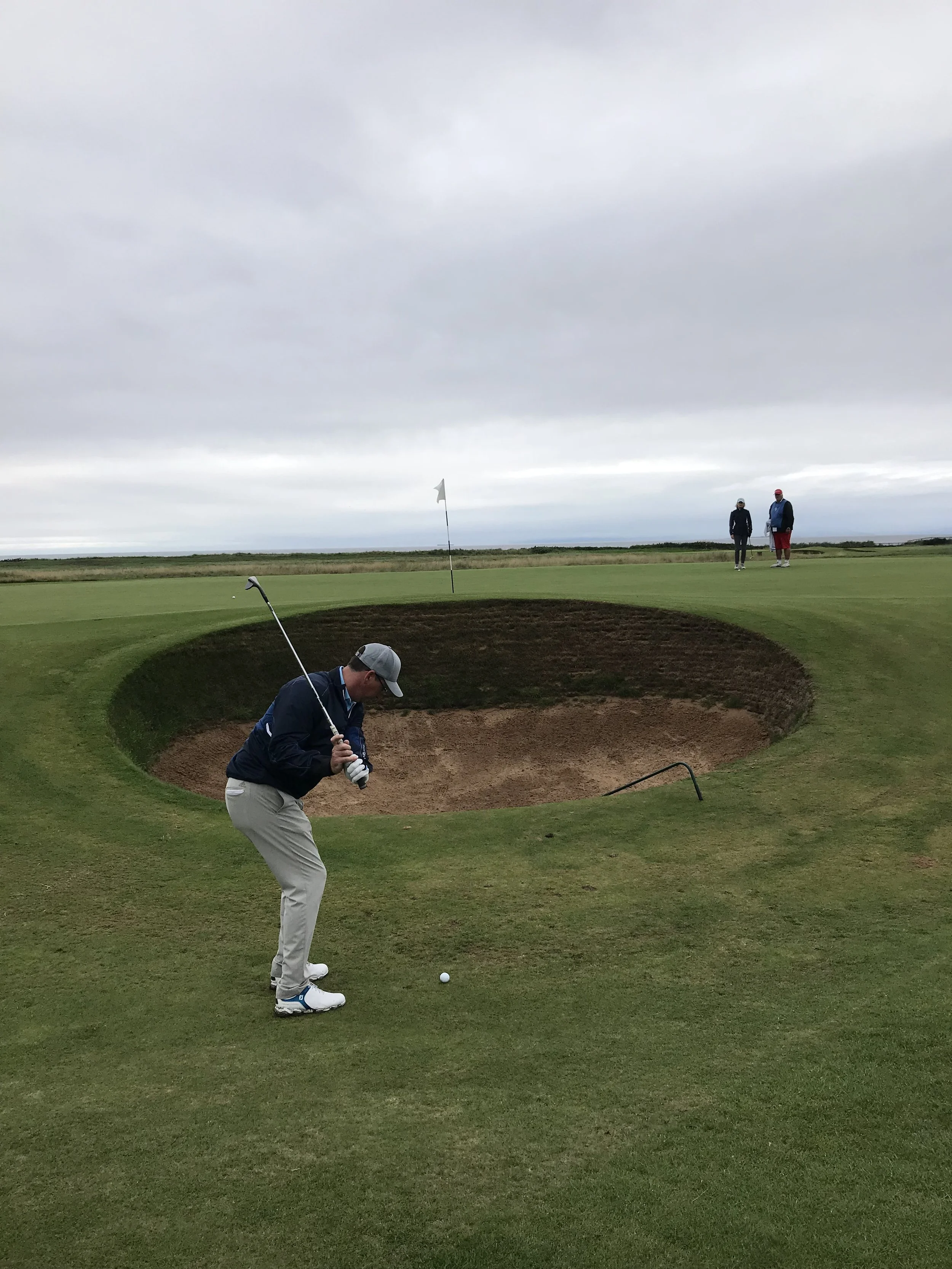 Golfer preparing to take a shot from a bunker on a golf course, with two people in the background standing near the green under a cloudy sky.