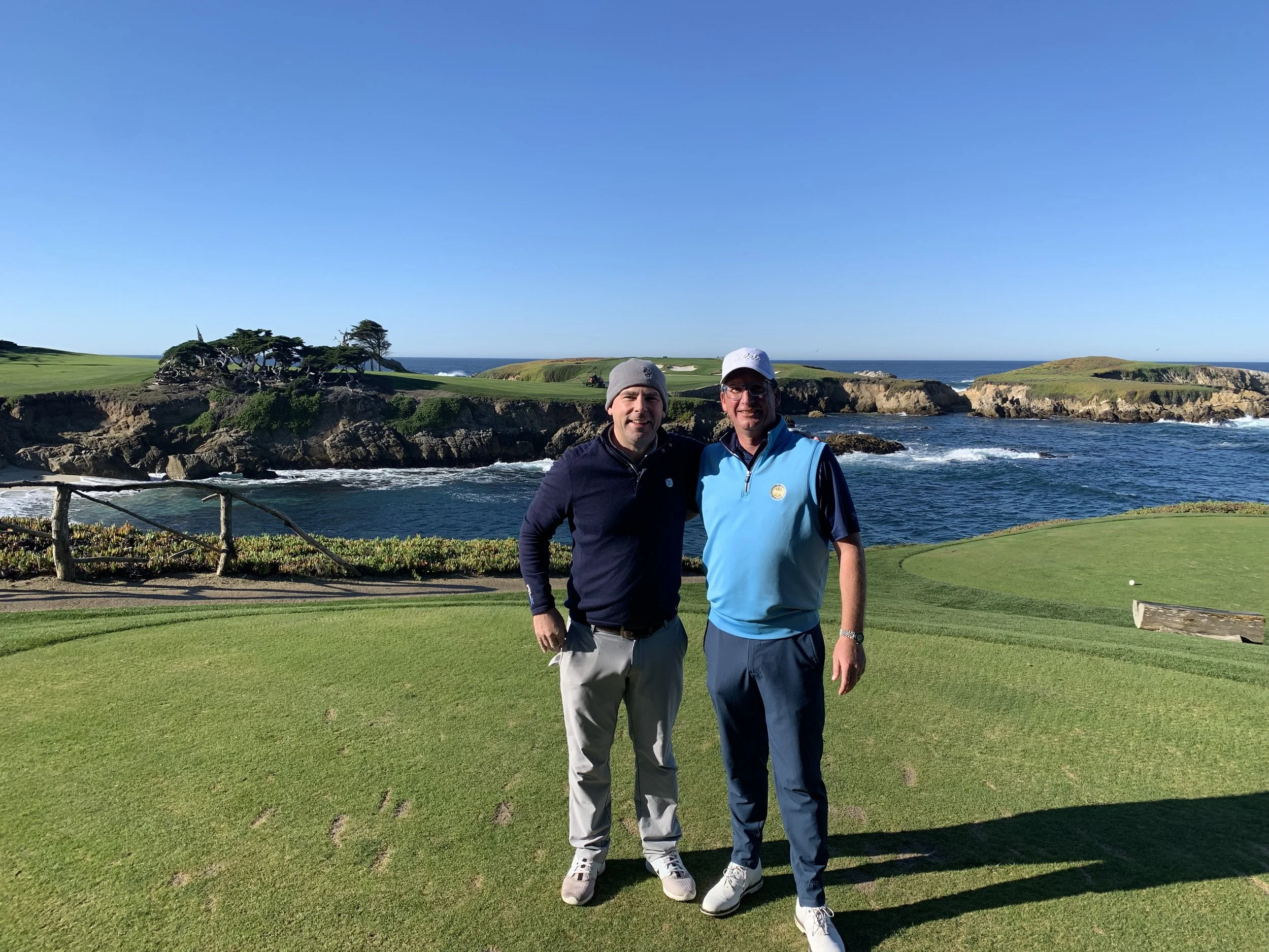Two men standing on a golf course with the ocean and rocky shoreline in the background.