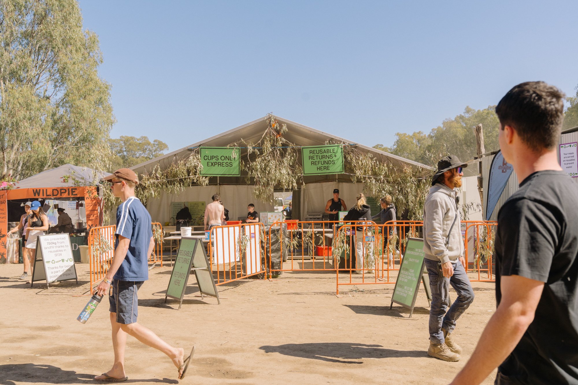 Outdoor event with a large white tent, surrounded by orange barriers, and signs indicating a reusable cups and refunds station. Several people are walking around, some near the tent and others in front, with trees and blue sky in the background.