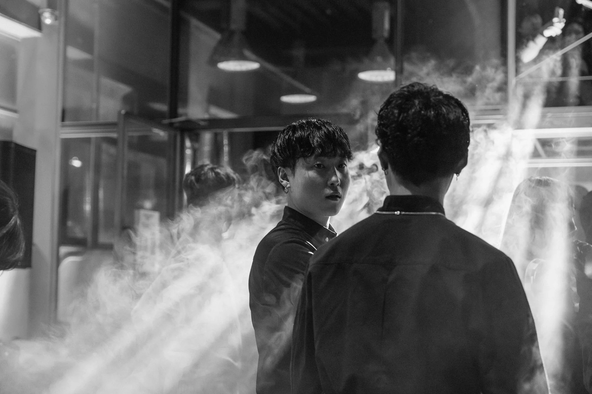 Two people having a conversation in a smoky, modern indoor setting, possibly a restaurant or café, with other people in the background.