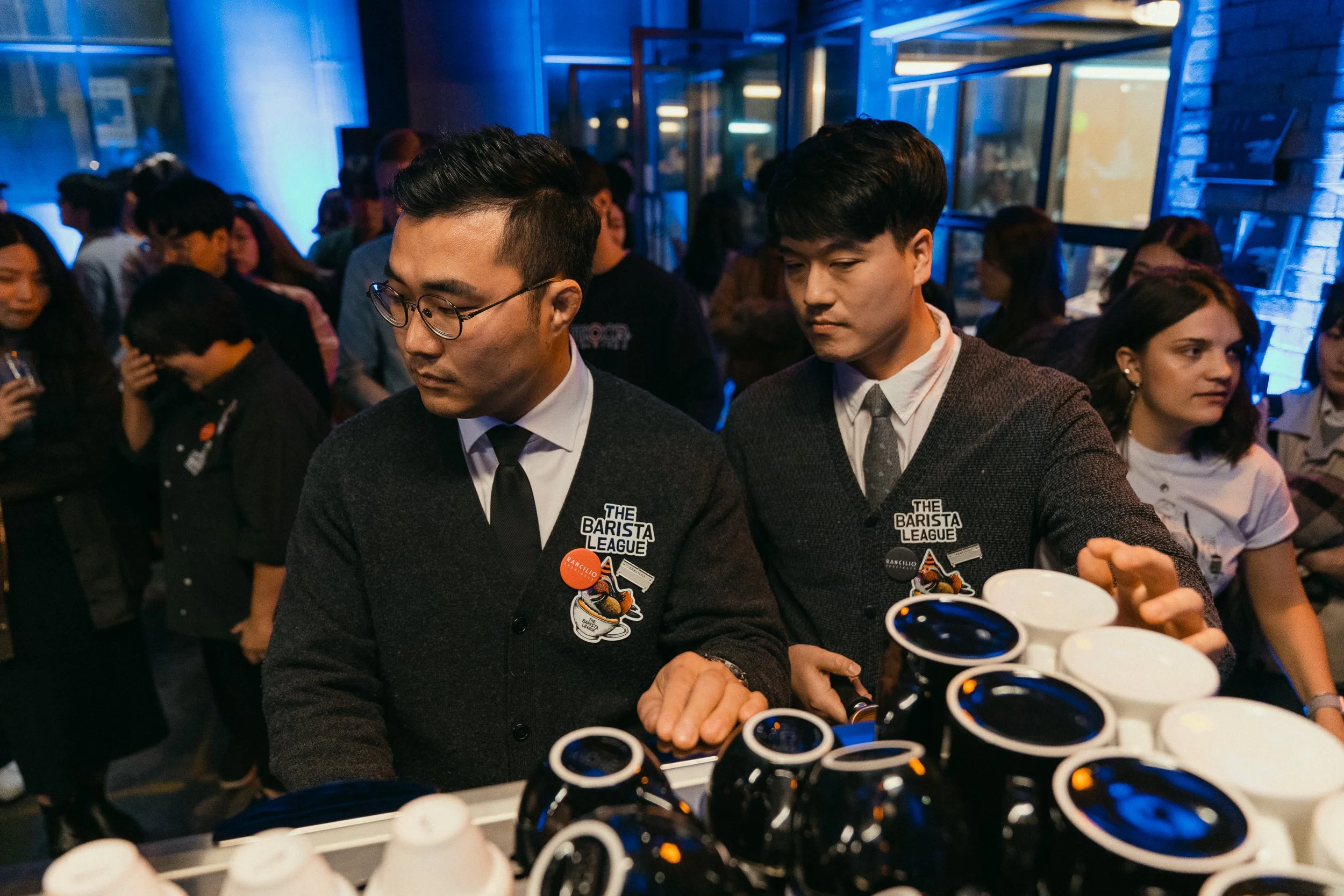 Two male baristas in dark sweaters with badges, preparing coffee in a busy cafe or event space, with a crowd of people in the background.