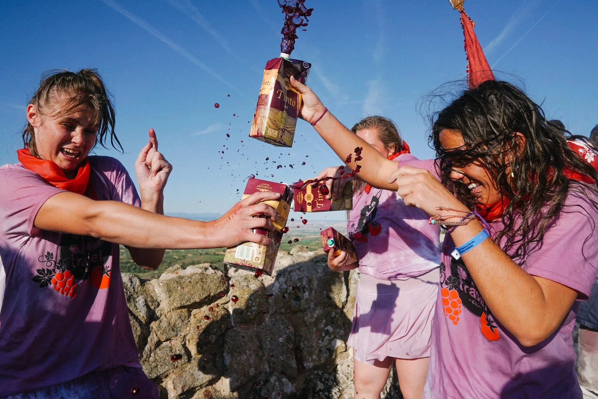 Group of young women in pink shirts celebrating outdoors, splashing cherry wine from cartons.
