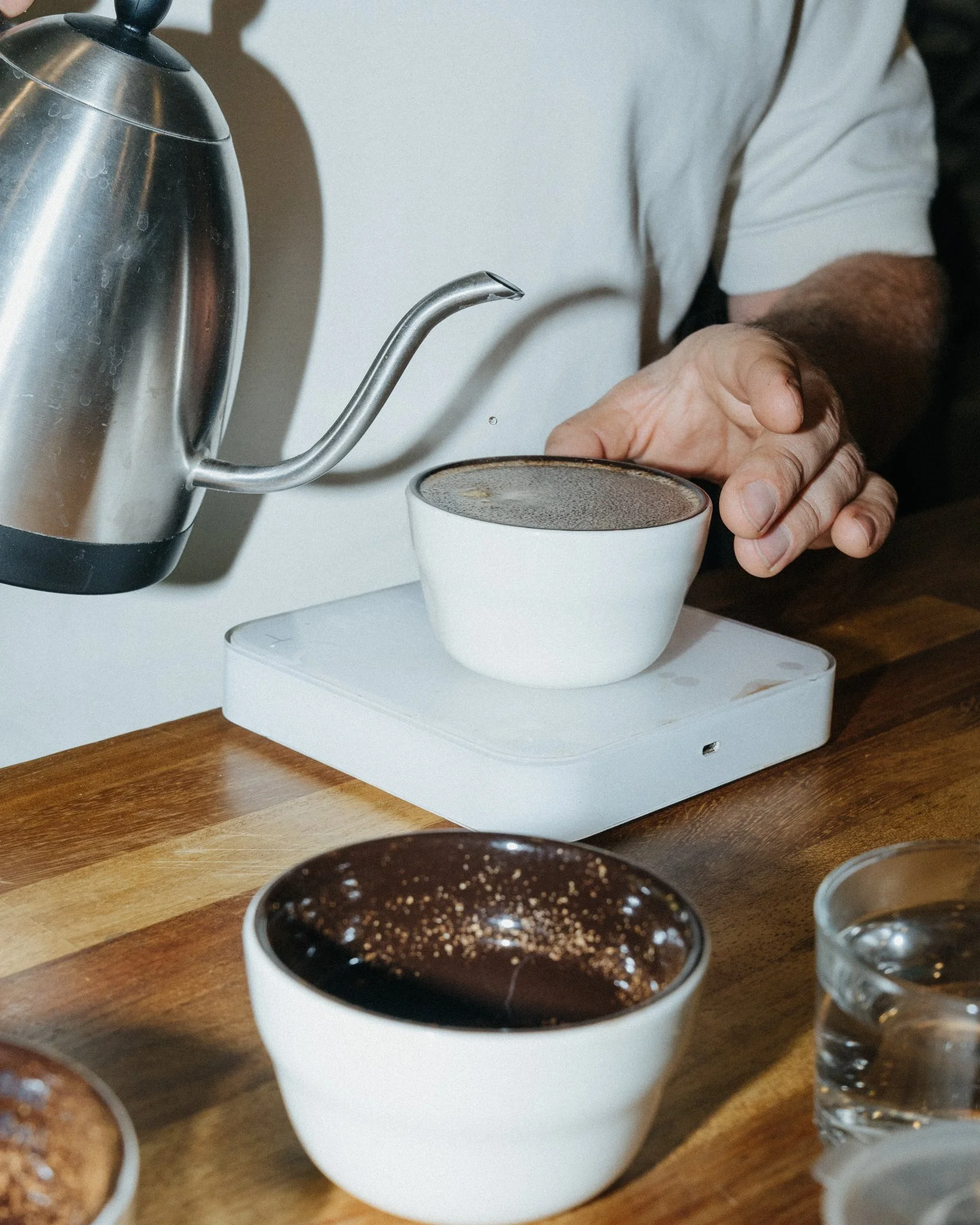 A person pouring hot water into a white bowl on a digital scale, with another bowl and a glass of water nearby on a wooden table.