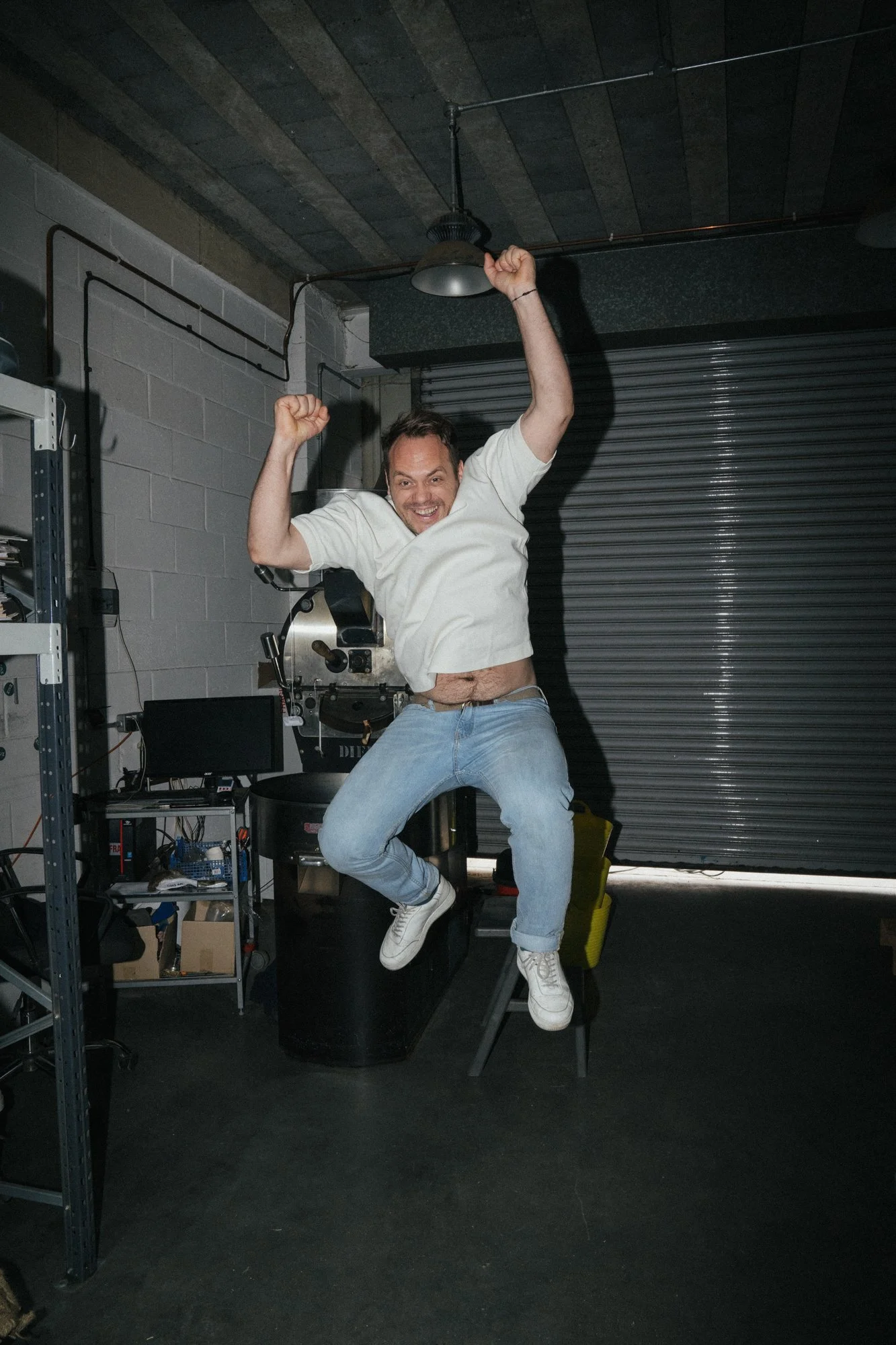 A man is jumping in the air with a big smile, arms raised in excitement, in an industrial-style room with a garage door, equipment, and a computer.