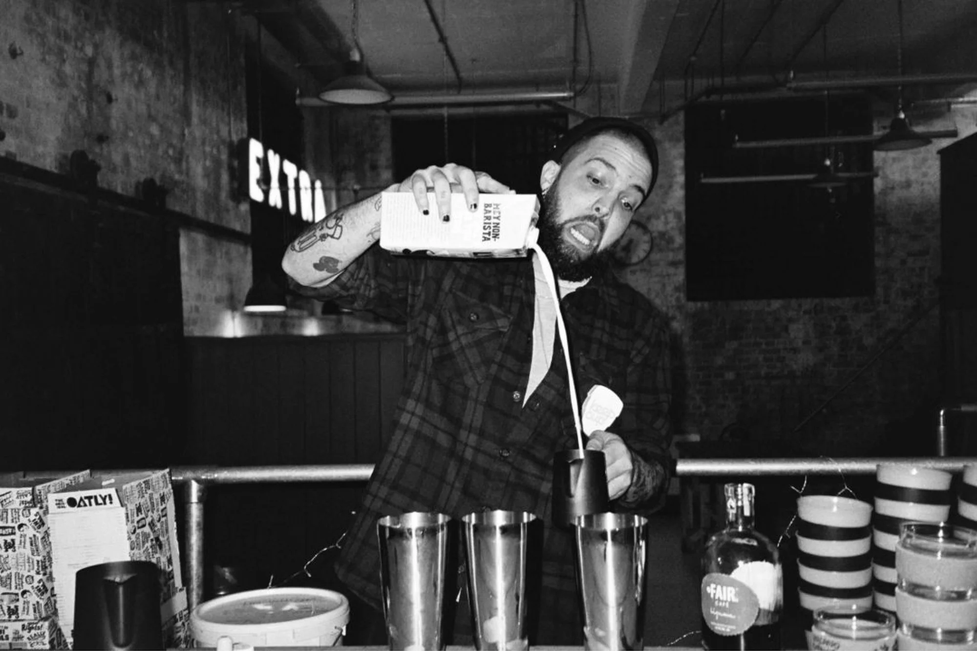 A man with tattoos pouring a drink from a carton into a black mug in a dimly lit bar or restaurant with exposed brick walls and an exit sign in the background.
