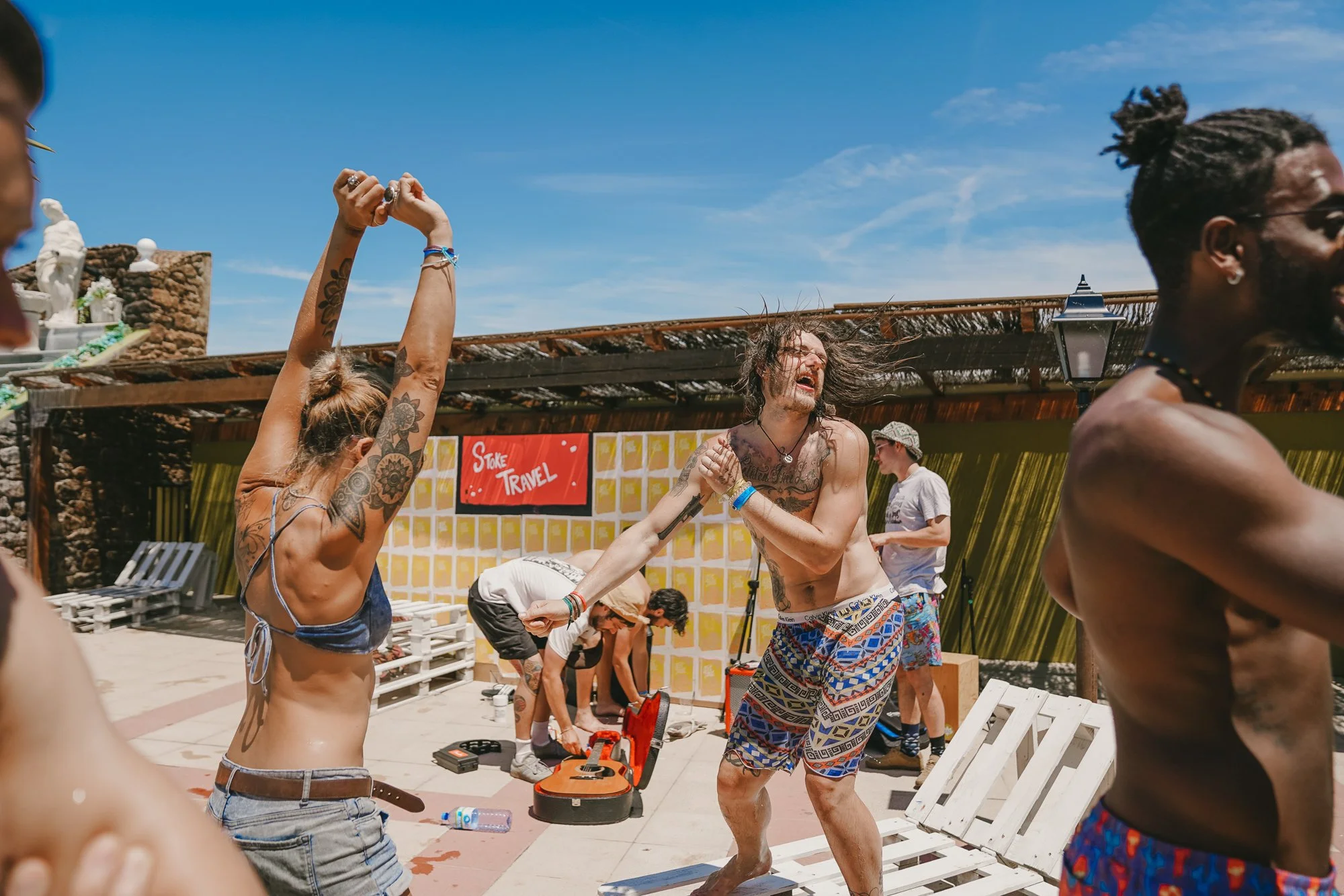 People dancing and enjoying themselves by a poolside, with one man playing guitar and others dancing under a bright blue sky at a daytime outdoor party.