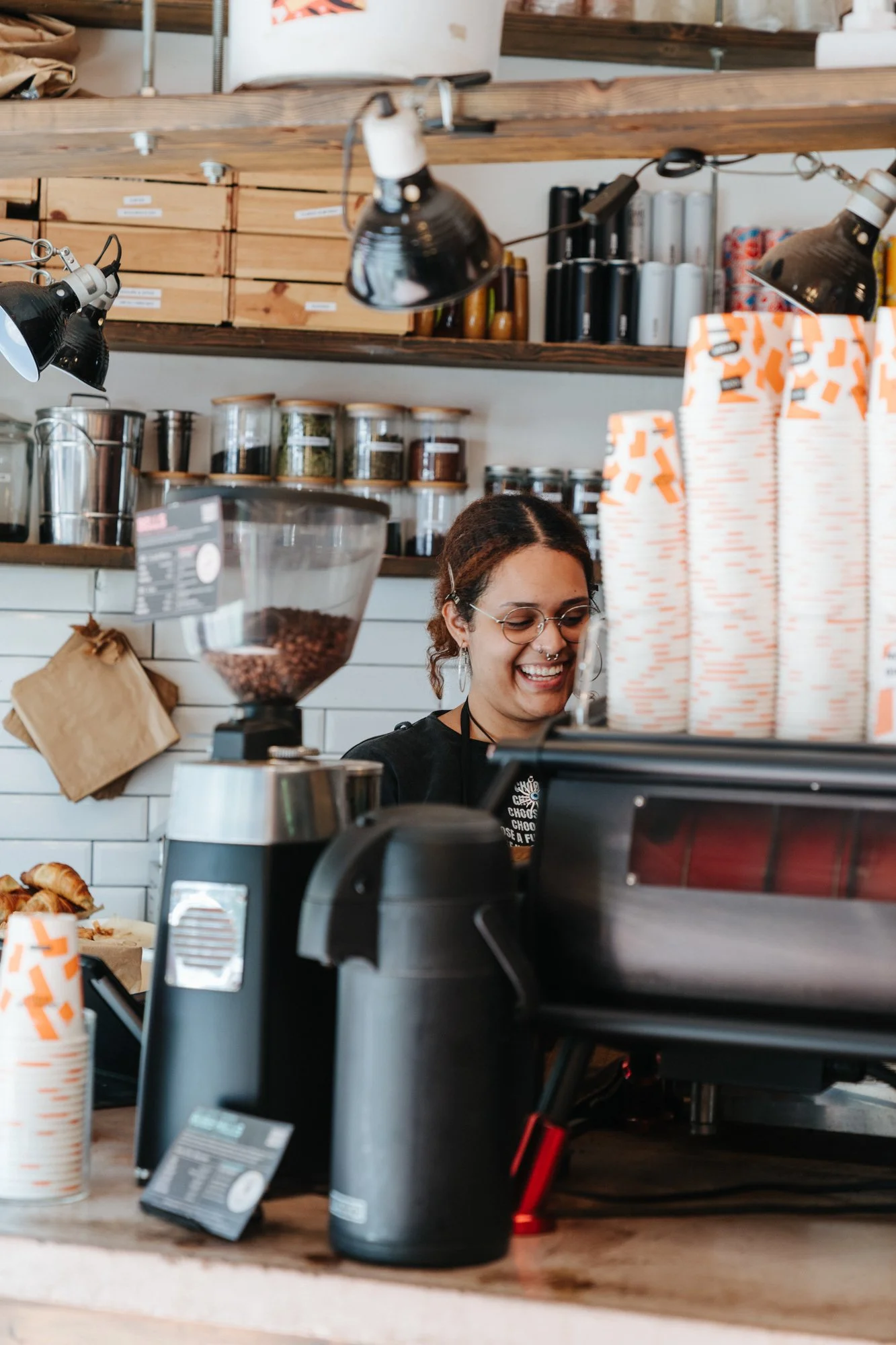 A woman working at a coffee shop, smiling behind the counter with various coffee-making equipment and supplies around her.