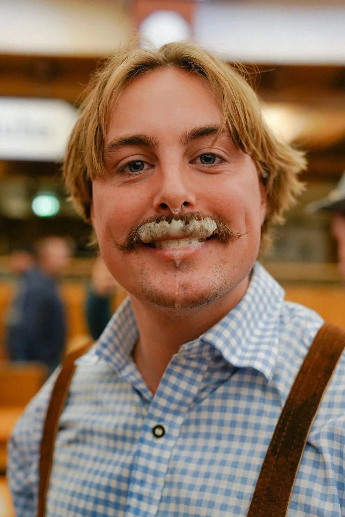 A young man with blonde hair, a mustache, and a goatee, smiling at the camera, wearing a blue gingham shirt and suspenders, in a rustic indoor setting.