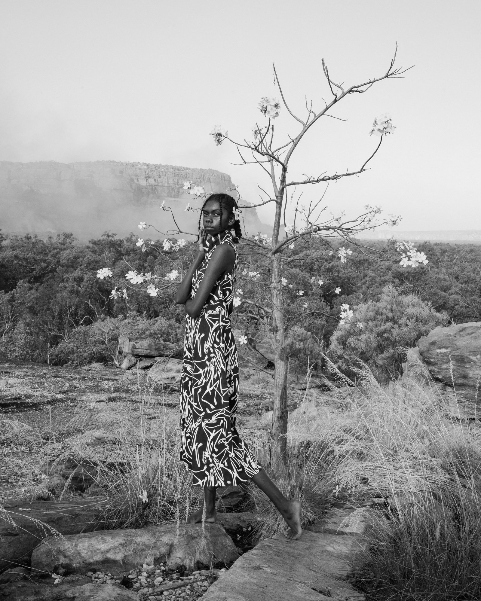 A black and white photo of a woman standing barefoot on rocks next to a small tree, with a distant landscape and cliffs in the background.