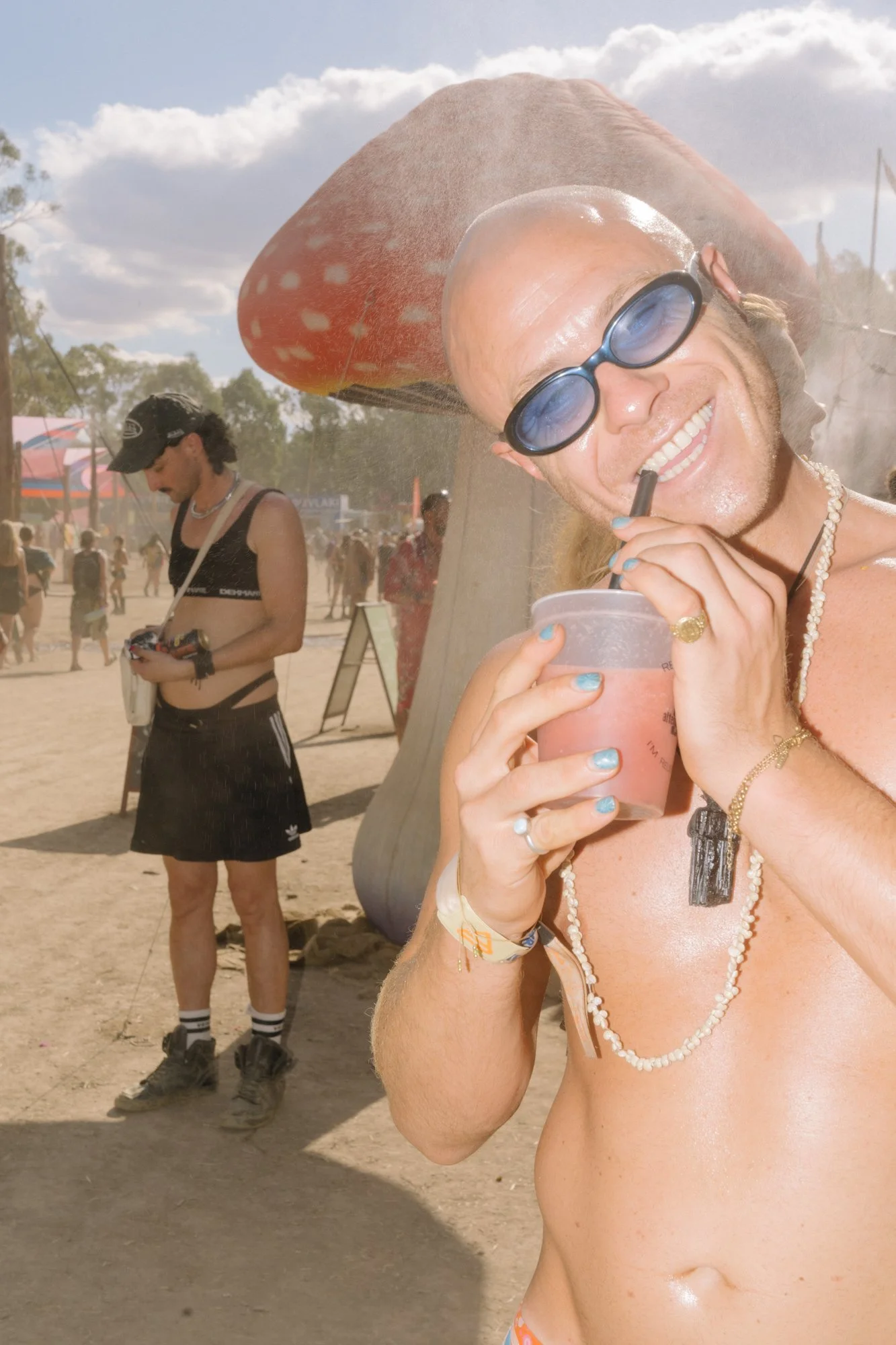 A man with long hair, sunglasses, and jewelry smiling and holding a pink drink with a straw at an outdoor festival. A large mushroom-shaped sculpture or structure is in the background, along with other festival attendees and trees.