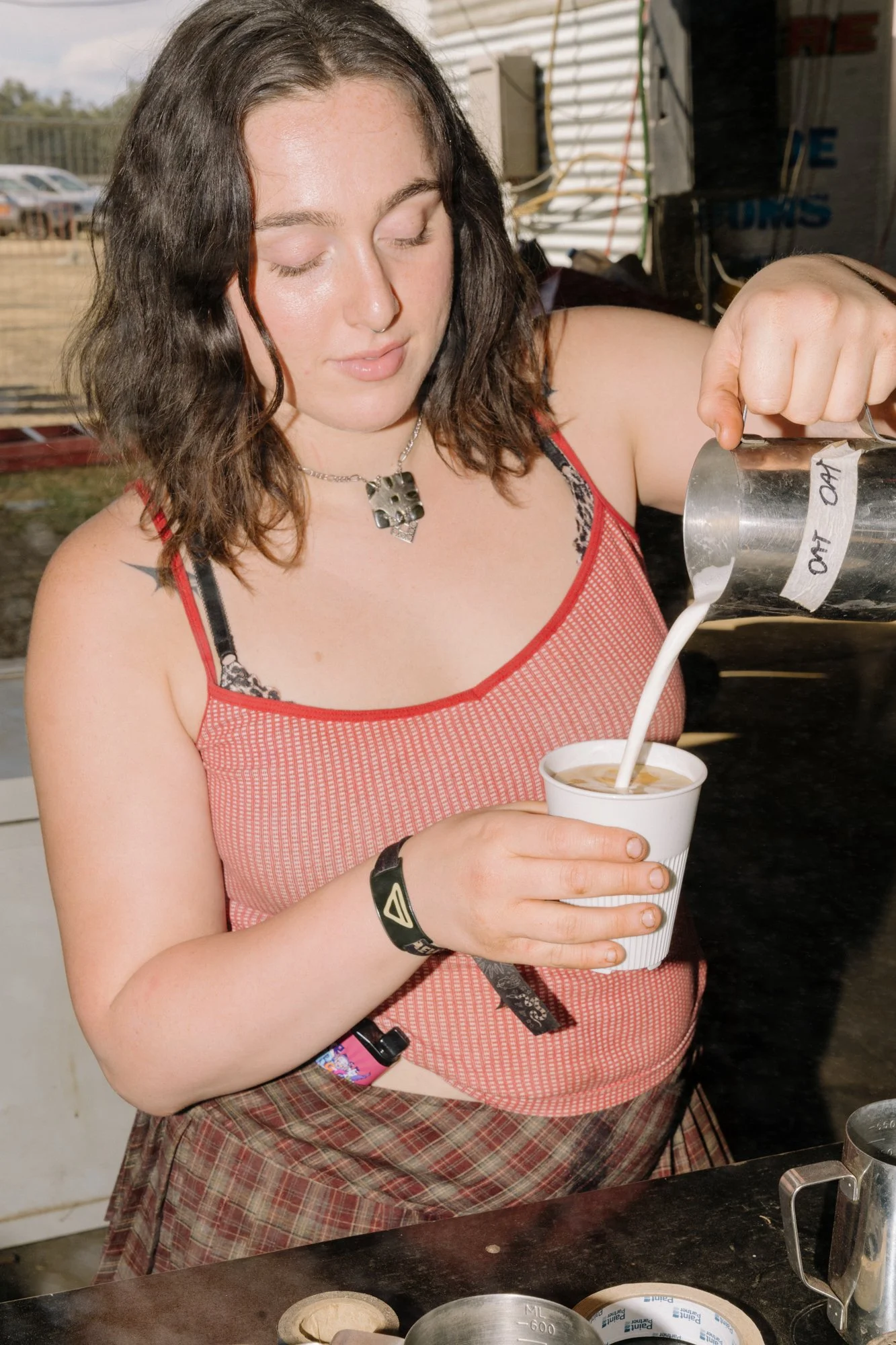 A woman with dark brown wavy hair, wearing a red tank top and plaid skirt, is pouring milk from a metal pitcher into a paper cup, with her eyes closed.