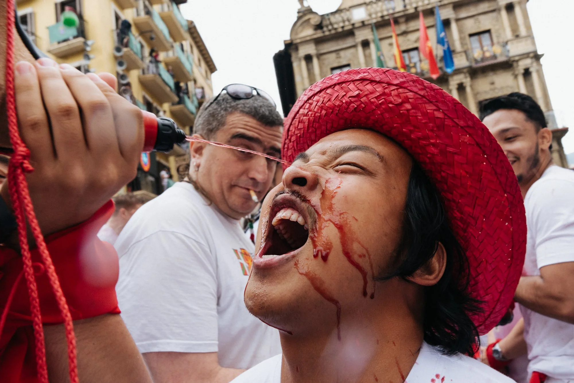 A woman wearing a red hat getting blood splattered on her face at a public event with other individuals and historic buildings in the background.